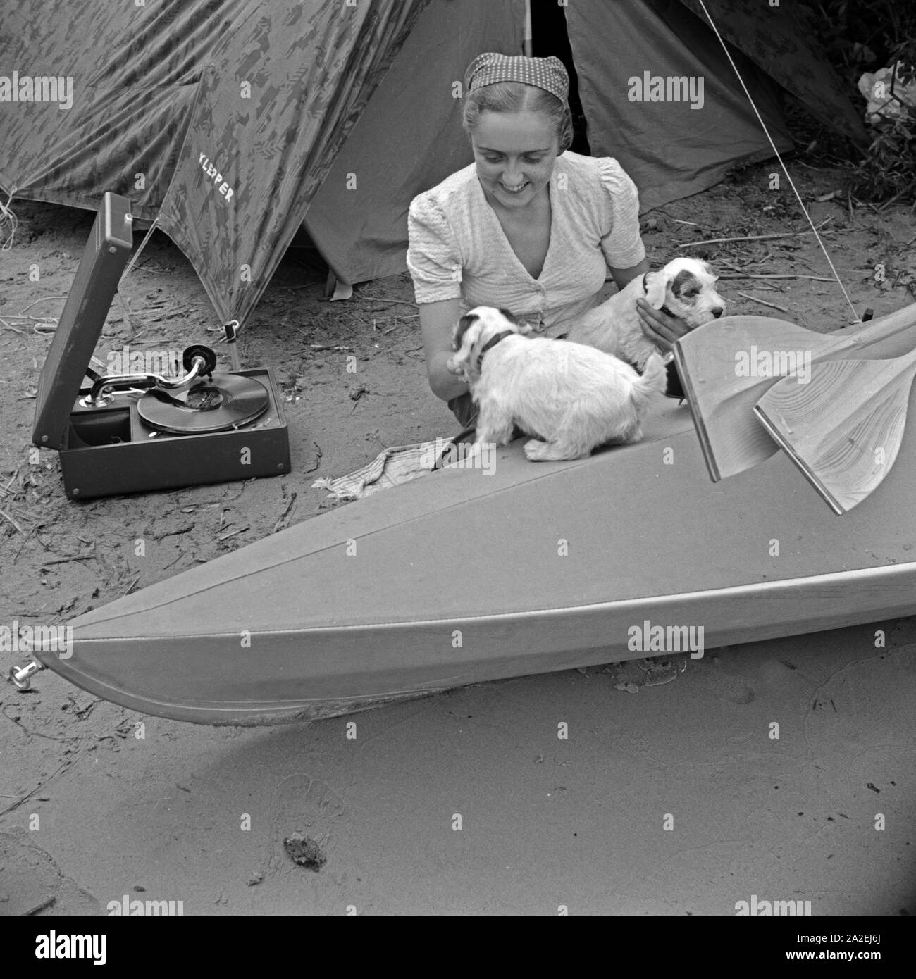 Werbefoto für KLEPPER Faltboot und Zelt: eine junge Frau spielt mit zwei Welpen vor einem Zelt und einem Boot, Deutschland 1930er Jahre. Werbung für ein Klepper foldboat und Zelt: Eine junge Frau spielen mit zwei Welpen vor einem Boot und einem Zelt am Ufer eines Sees, Deutschland 1930. Stockfoto