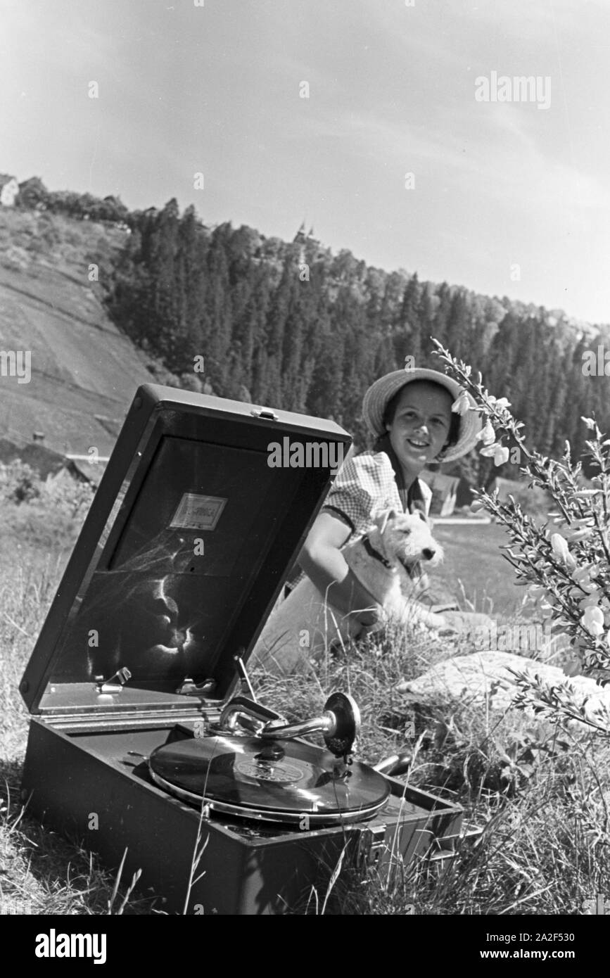 Eine junge Frau hört sich eine Platte der Schallplattenfirma Electrola mit dem Koffergrammophon auf der Wiese ein, Deutschland 1930er Jahre. Eine junge Frau, die Wiedergabe einer Aufzeichnung der Plattenfirma Electrola auf einem tragbaren Wind spielte, Gramophone, Deutschland 1930. Stockfoto