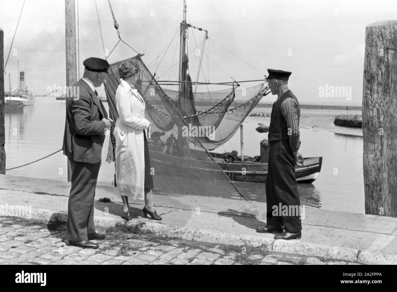 Eine Frau und ein Mann treffen einen Fischer vor seinem Fischerboot in Norddeich, Deutschland 1930er Jahre. Eine Frau und ein Mann treffen ein Fischer vor seinem Boot bei Norddeich, Deutschland 1930. Stockfoto