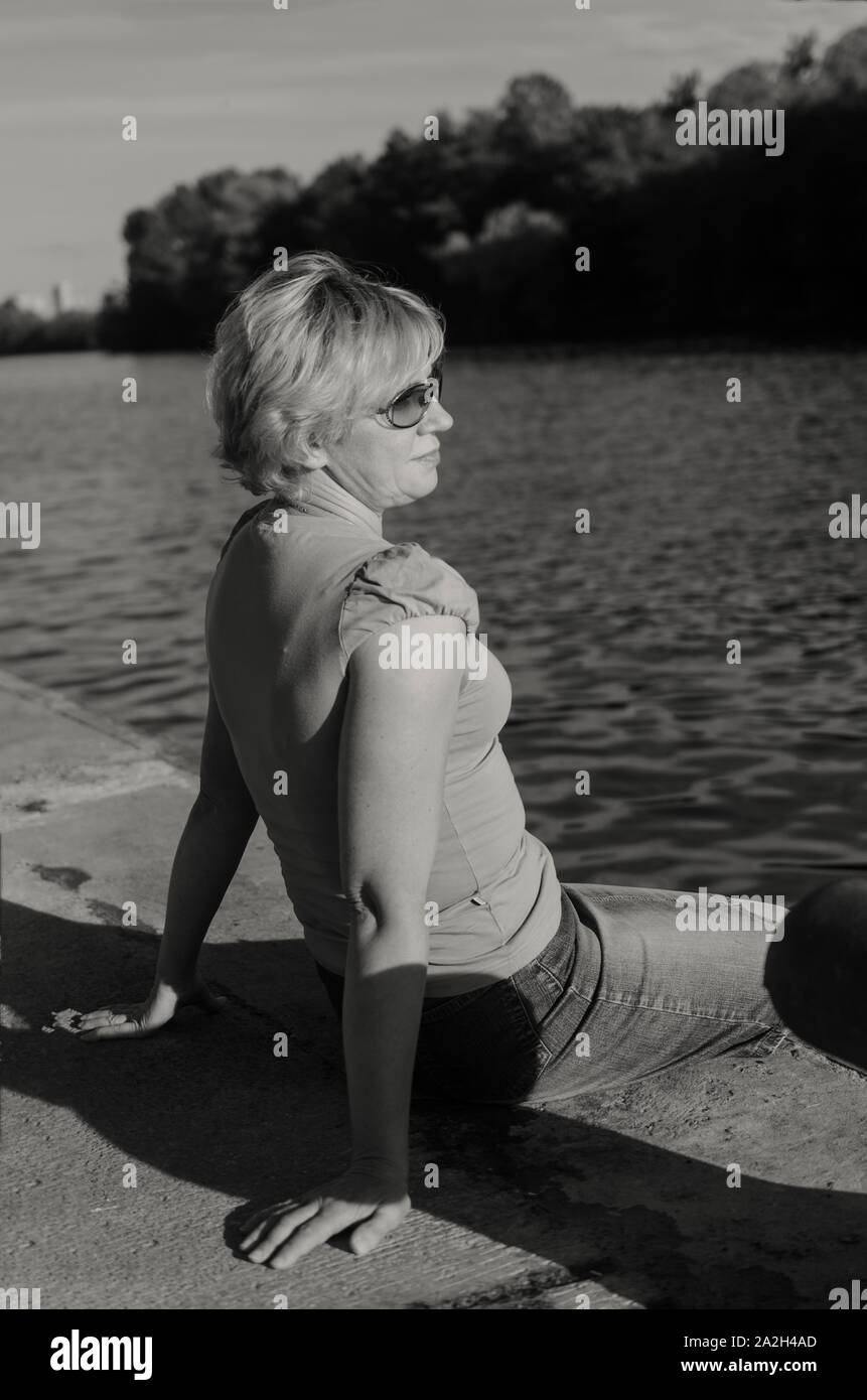 Eine Frau von 45-50 Jahre alt sitzt mit Traurigkeit in der Nähe des Flusses auf dem Damm Straße Stockfoto