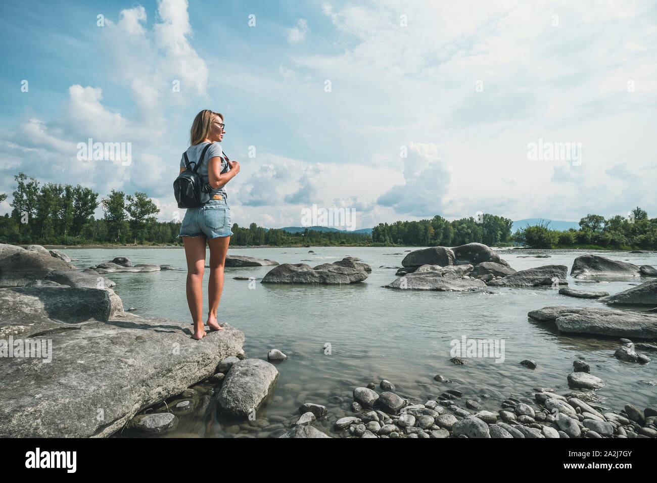 Stilvolle Blondine mit kurzen Hosen mit Rucksack in der Nähe von Berge, Fluss und Wald. Blondes Haar Frau Natur erkunden. Reisen und Fernweh Konzept. Amaz Stockfoto