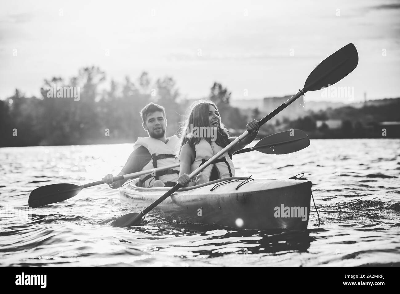 Glückliche Junge kaukasier Paar Kajakfahren auf dem Fluss mit Sonnenuntergang im Hintergrund. Spaß in der Freizeit. Gerne männliche und weibliche Modell laughting auf dem Kajak. Sport, Beziehungen Konzept. Farblos. Stockfoto