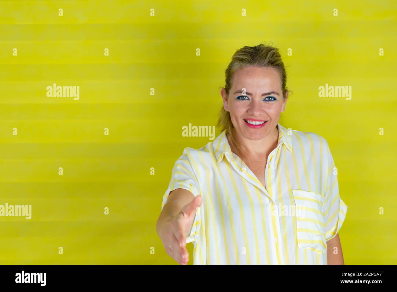 Junge schöne Frau mit gestreiften Hemd stehend über isolierte gelben Hintergrund lächelt freundlich mit Handshake als Gruß und einladend. Stockfoto