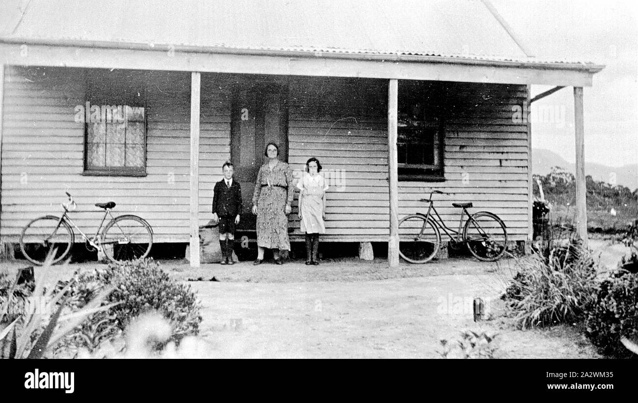 Negative - Familie Gruppe & Fahrräder außerhalb Farm House, Mooralla, Victoria, ca. 1935, eine Frau, ein kleiner Junge und ein Mädchen stehen vor einem Haus. Die Frau und das kleine Mädchen tragen Kleider, und der kleine Junge trägt eine Jacke mit Shorts. Es gibt zwei Fahrräder, eine für ein Mädchen, lehnte sich gegen das Haus. Das Haus verfügt über zwei große Fenster auf beiden Seiten der Tür, und die Vorhänge können durch das Fenster auf der linken Seite gesehen werden. Das Haus ist im viktorianischen Stil Holz mit einem Dach Stockfoto