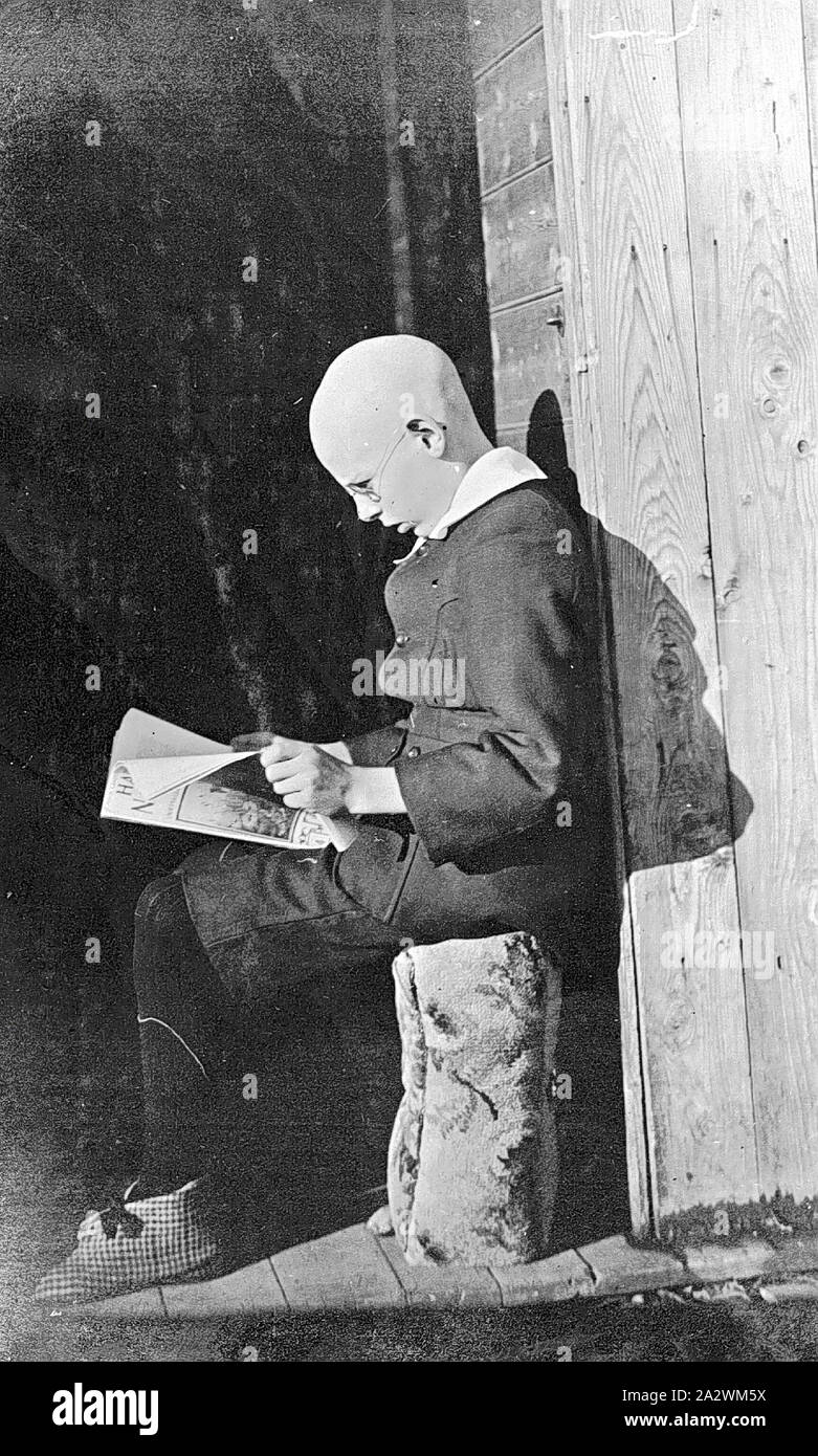 Negative sitzende Junge Lesen einer Zeitschrift, Merrigum, Victoria, 1910, ein Junge, mit kahl rasierten Kopf und elegant gekleidet, Sitzen beim Lesen einer Zeitschrift Stockfoto