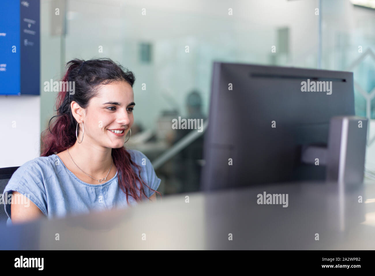 Junge Frau lächelnd beobachten, Nachrichten auf dem Bildschirm in einem Coworking Space. Moderne Einrichtung mit Glas Divisionen für freiberufliche Mitarbeiter. Stockfoto