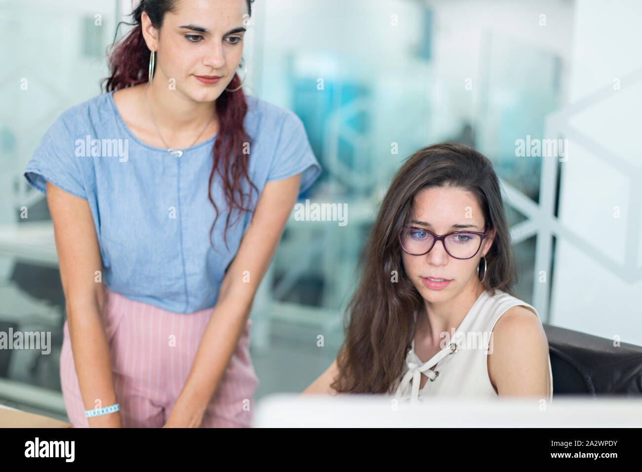 Zwei junge Frauen beobachten, Nachrichten auf dem Bildschirm in einem Coworking Space. Moderne Einrichtung mit Glas Divisionen für freiberufliche Mitarbeiter. Stockfoto