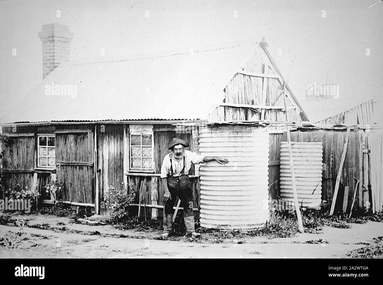 Negative - Bairnsdale Bezirk, Victoria, ca. 1890, ein älterer Mann außerhalb einer Bark Hut mit einem Wellblechdach. Er steht mit seiner Hand auf einem von zwei Wassertanks Stockfoto