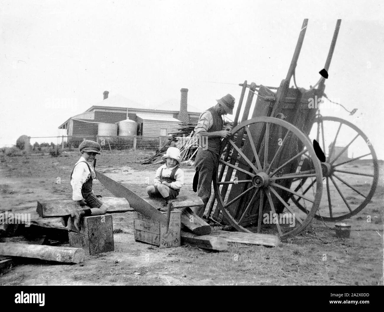 Negative - Sandford Bezirk, Victoria, 1927, ein Mann der Reparatur einer Feder Warenkorb. Zwei kleine Jungen sitzen auf ein Archiv, in dem ein Quersäge eingebettet ist. Das 'One Tree Hill' Homestead ist im Hintergrund Stockfoto