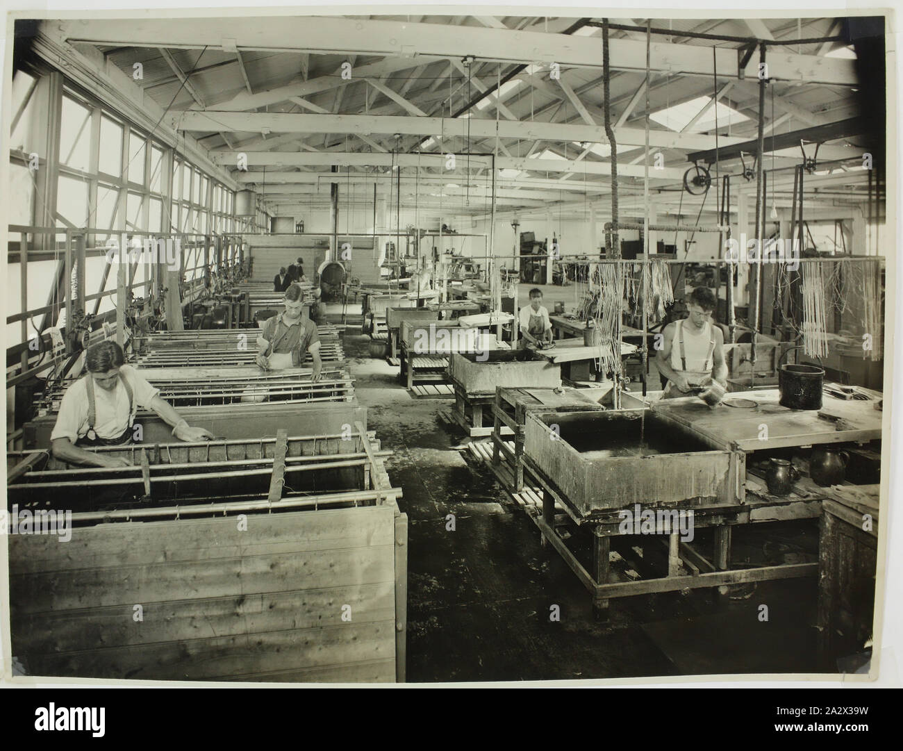 Fotografie - Hecla Elektrik Pty Ltd, Arbeiter, in der Galvanik, ca. 1925, schwarz-weiß Foto von männlichen Fabrikarbeiter plating Gerät bei Hecla Werk, wahrscheinlich der Little Bourke Street. An der linken Wand Komponenten sind in Wannen von Lösung für die Galvanotechnik eingetaucht. nach rechts, andere Arbeitnehmer Teile reinigen. Die Gegend ist beleuchtet, die von Windows, die über der linken Wand, diese geöffnet sind, Lüftung. Ein Arbeiter Stockfoto