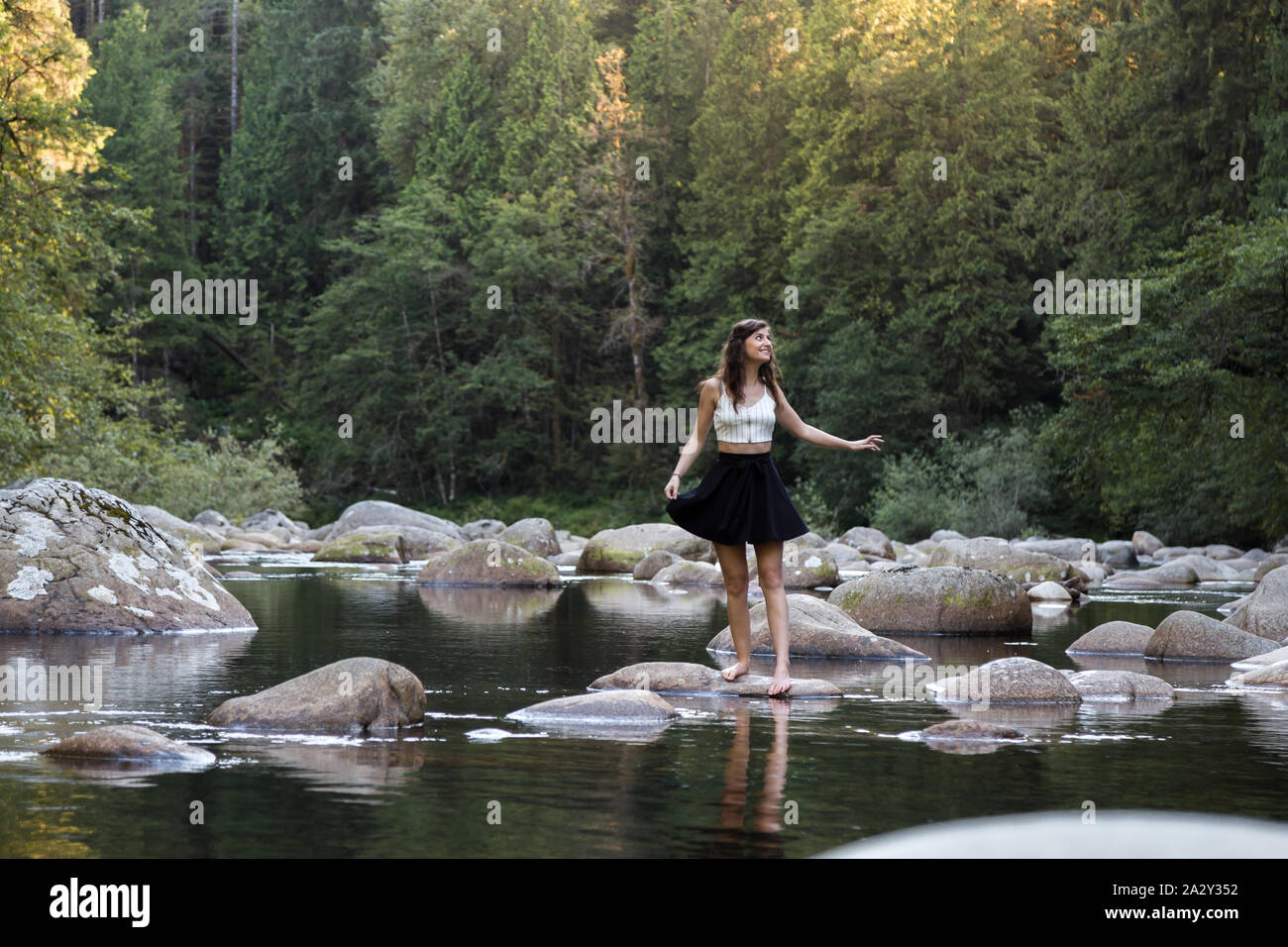 Junge attraktive Brünette Frau auf einem Felsen neben einem unberührten Fluss in einem immergrünen Wald. Stockfoto