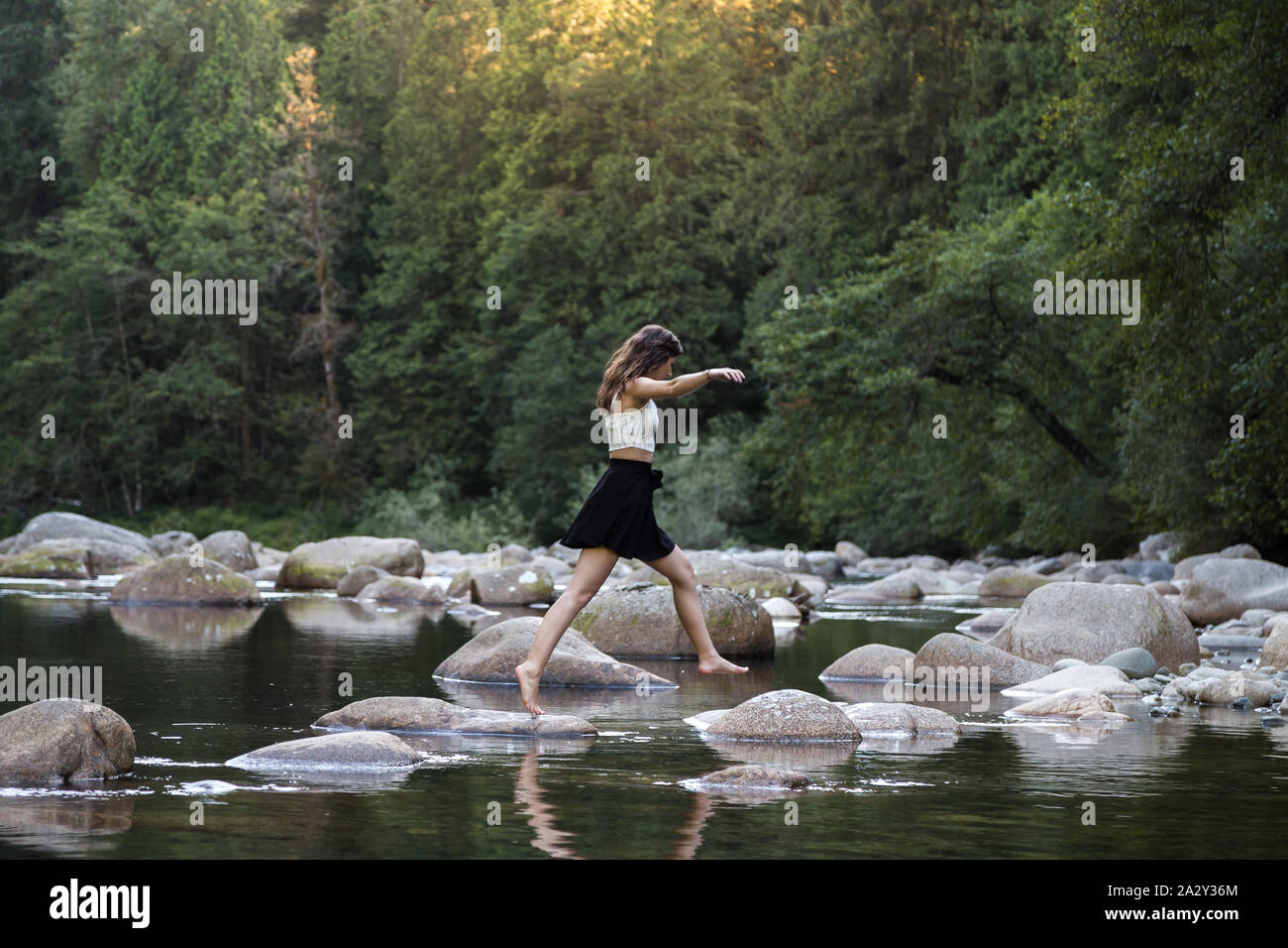 Junge attraktive Brünette Frau hüpft über Felsen neben einem unberührten Fluss in einem immergrünen Wald. Stockfoto