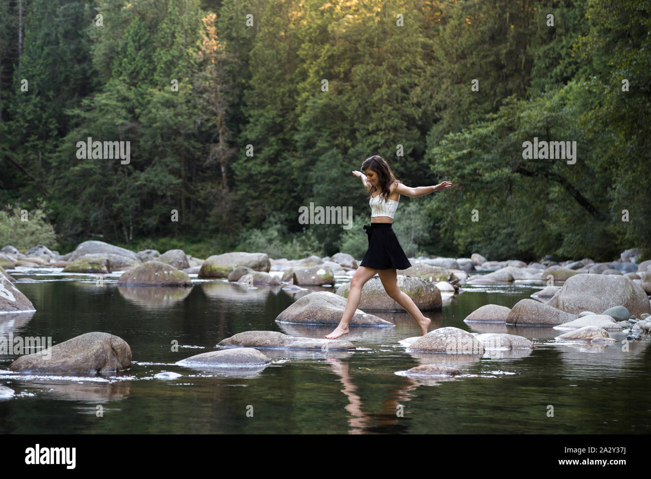 Junge attraktive Brünette Frau hüpft über Felsen neben einem unberührten Fluss in einem immergrünen Wald. Stockfoto