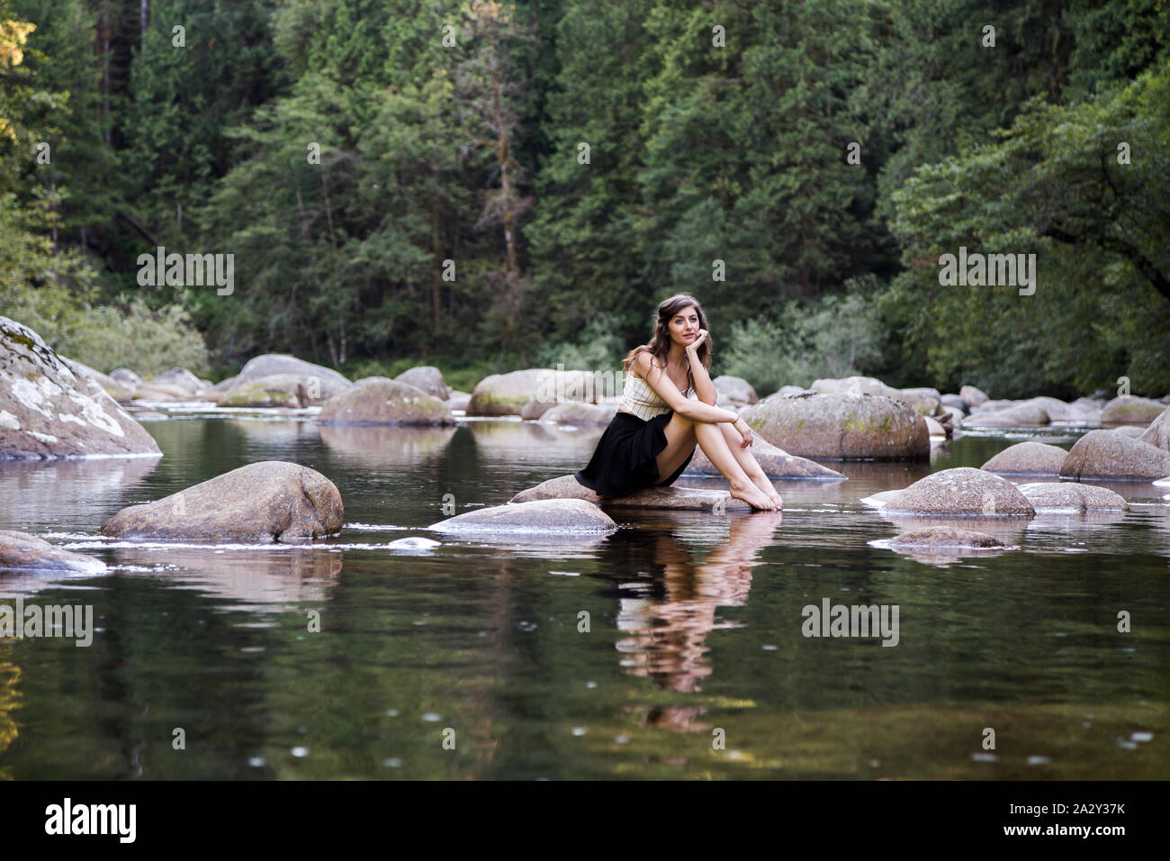 Junge attraktive Brünette Frau auf einem Felsen neben einem unberührten Fluss in einem immergrünen Wald. Stockfoto