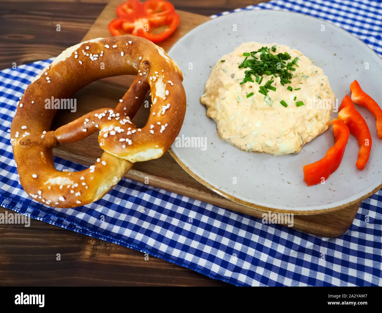 Traditionell bayerische Speisen Vorspeise mit Brezen und Käse Paste namens obazda auf Blau-weiß karierte Tischdecke und hölzerne Platte Stockfoto