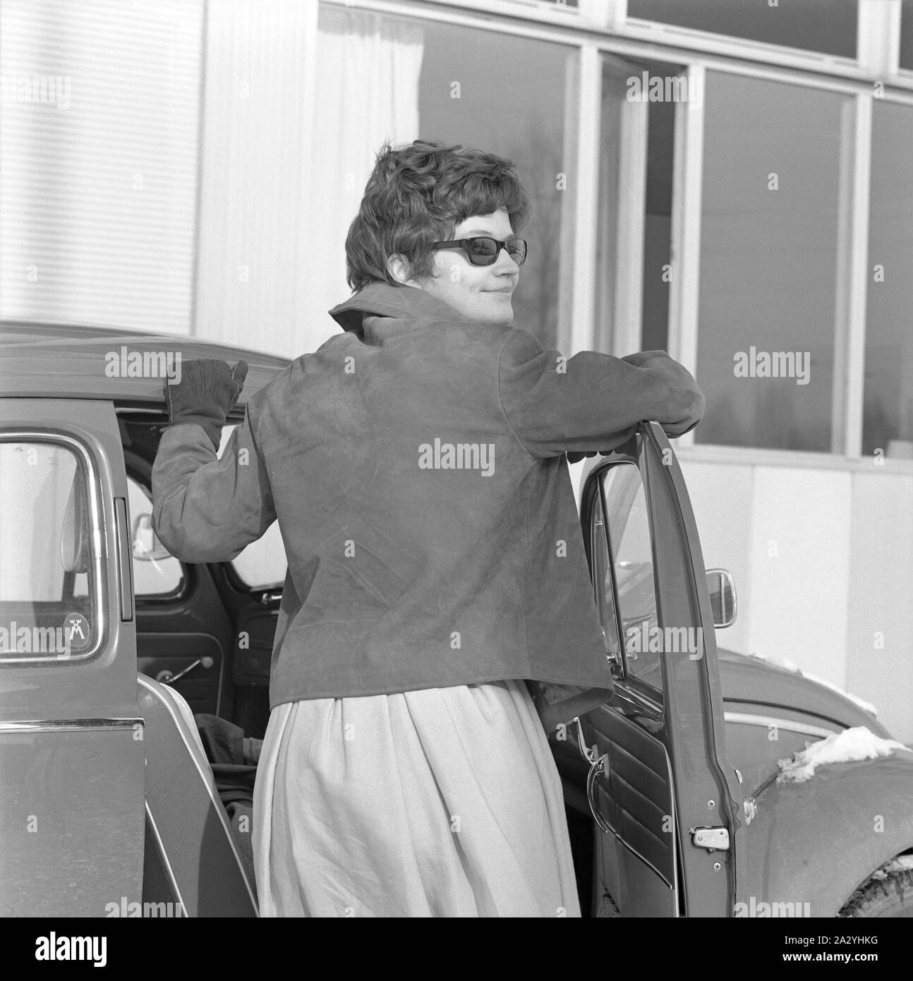 Das Fahren in den 1960er Jahren. Eine Frau, die an Ihrem Auto ein VW Käfer. Sie trägt einen Rock, eine kurze Lederjacke, Sonnenbrille und Handschuhe. Schweden 1962 Stockfoto
