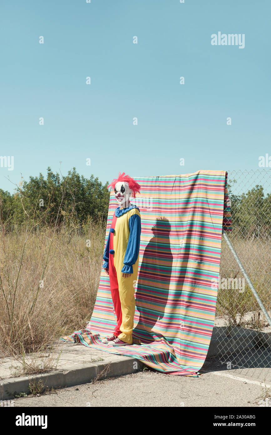 Ein furchtsames Clown in farbenfroher, gelben, roten und blauen Kostüm, vor einem Zaun, mit farbenfrohen, gestreiften Tuch hängen von ihr, und ein Stockfoto