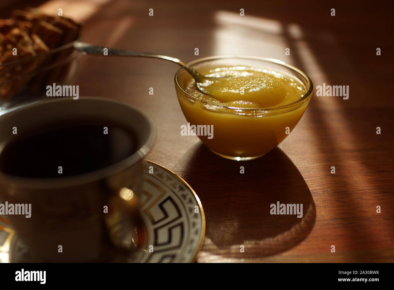Honig in einem Glas Schale mit einem Löffel, eine Tasse Kaffee auf einer Untertasse, Cookies in einer Vase auf der sonnigen Tabelle. Stockfoto