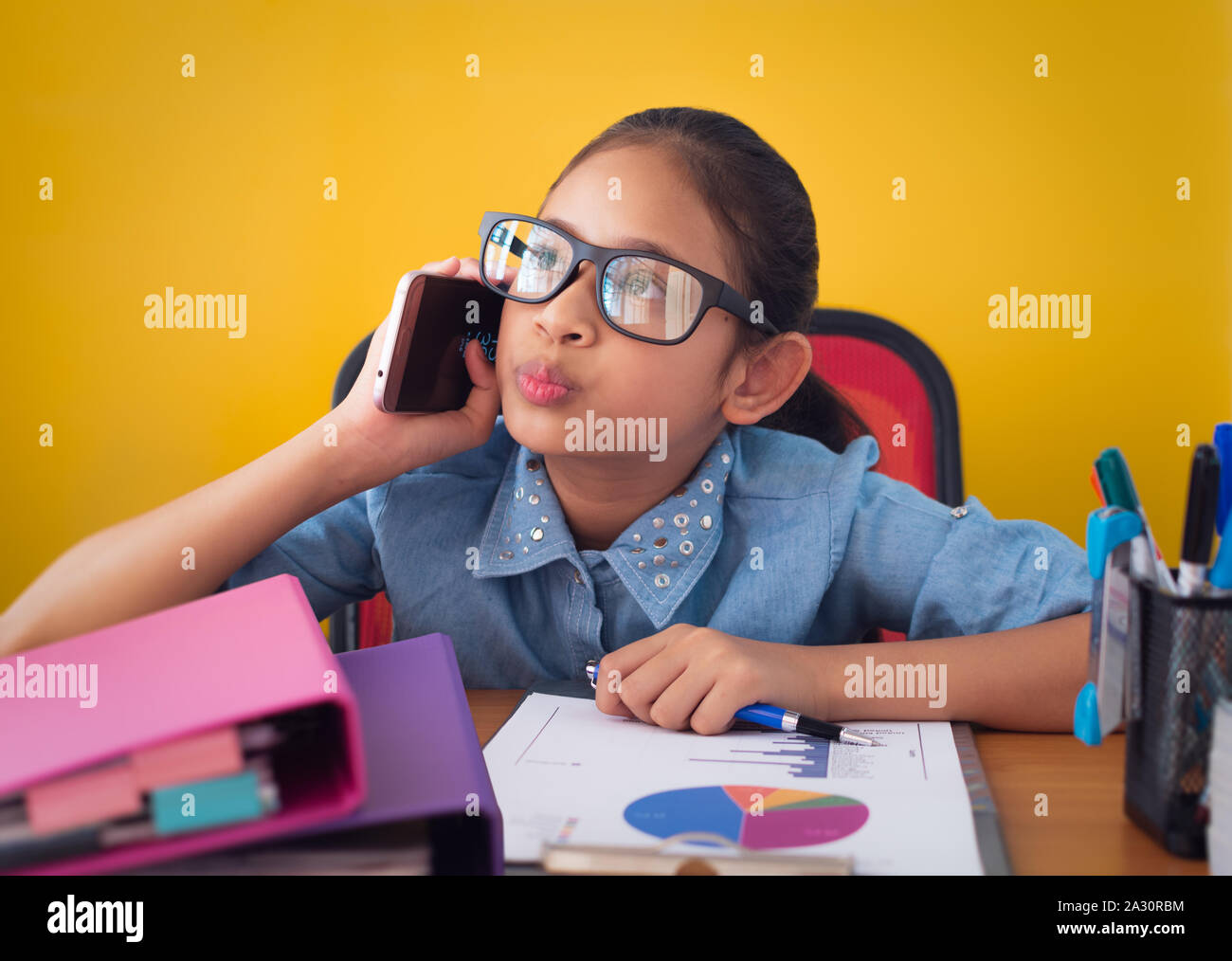 Süße Mädchen denken und sprechen über das Telefon am Schreibtisch isoliert gelben Hintergrund, Bildung und Kommunikation Konzept. Stockfoto
