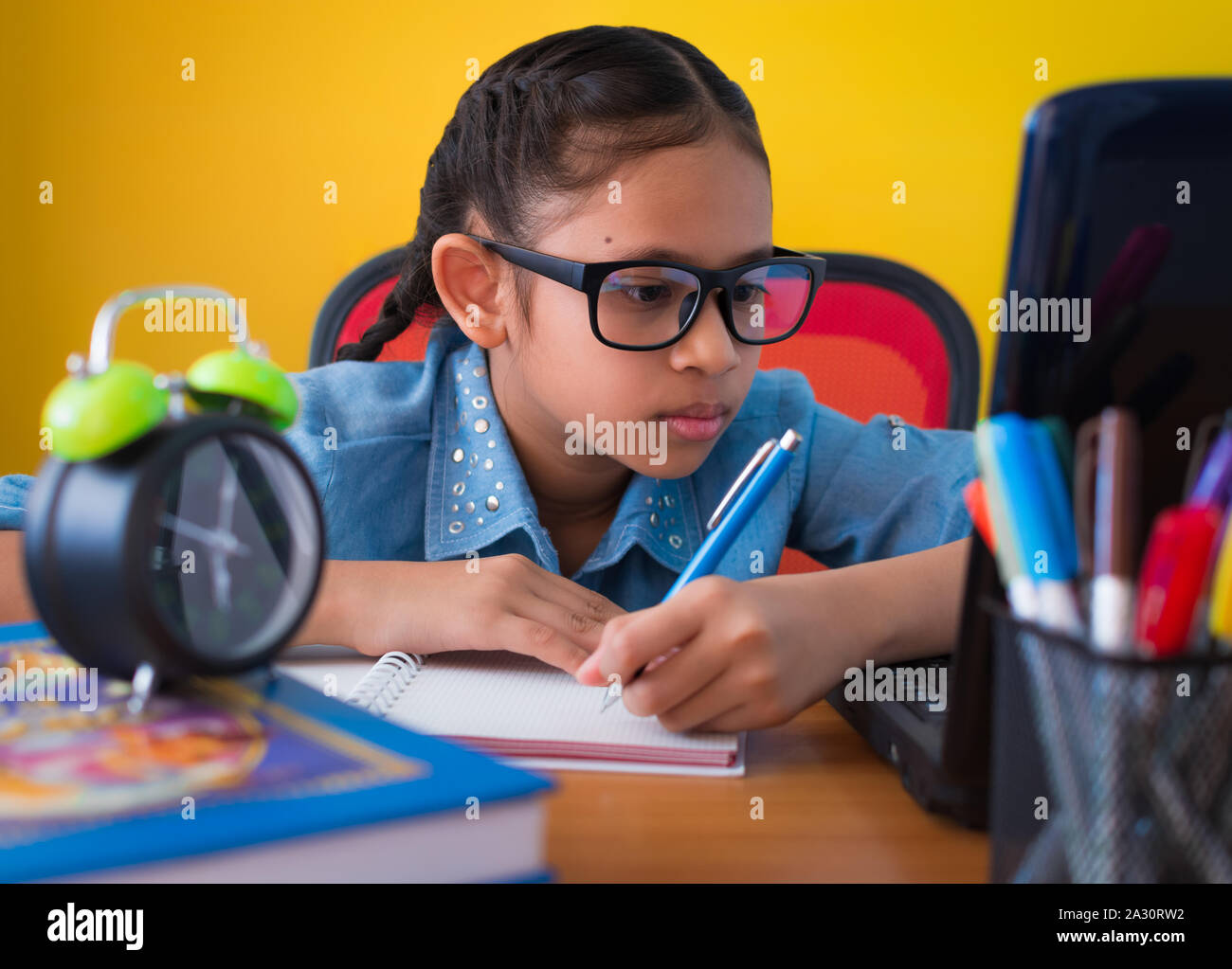 Nettes Mädchen mit Brille ist das Schreiben und mit Laptop auf dem Schreibtisch isoliert gelben Hintergrund, Bildung Konzept. Stockfoto