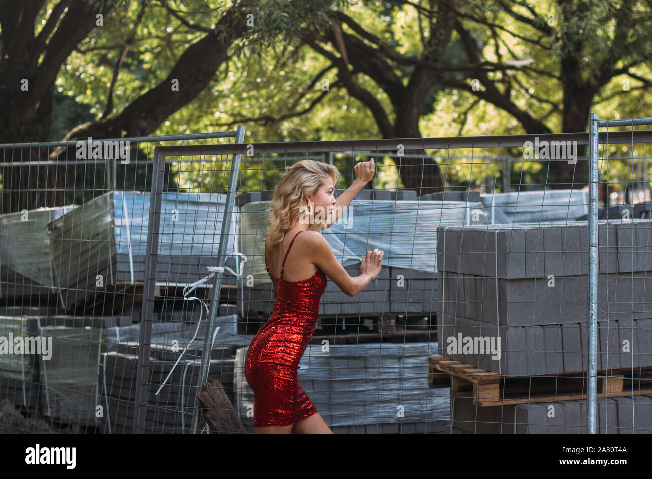 Schöne blonde Mädchen im roten Kleid mit Pailletten im Park von der Seite in der Nähe der Baustelle Stockfoto