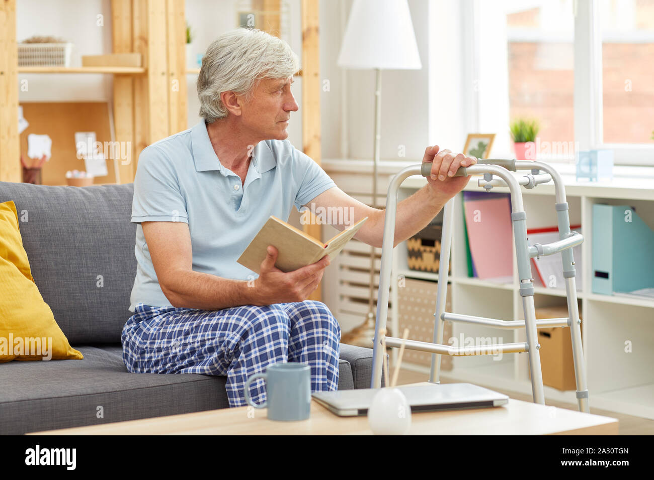 Ältere Menschen in Freizeitkleidung sitzt auf einem Sofa mit Walker und Er wird ein Buch zu Hause zu lesen Stockfoto