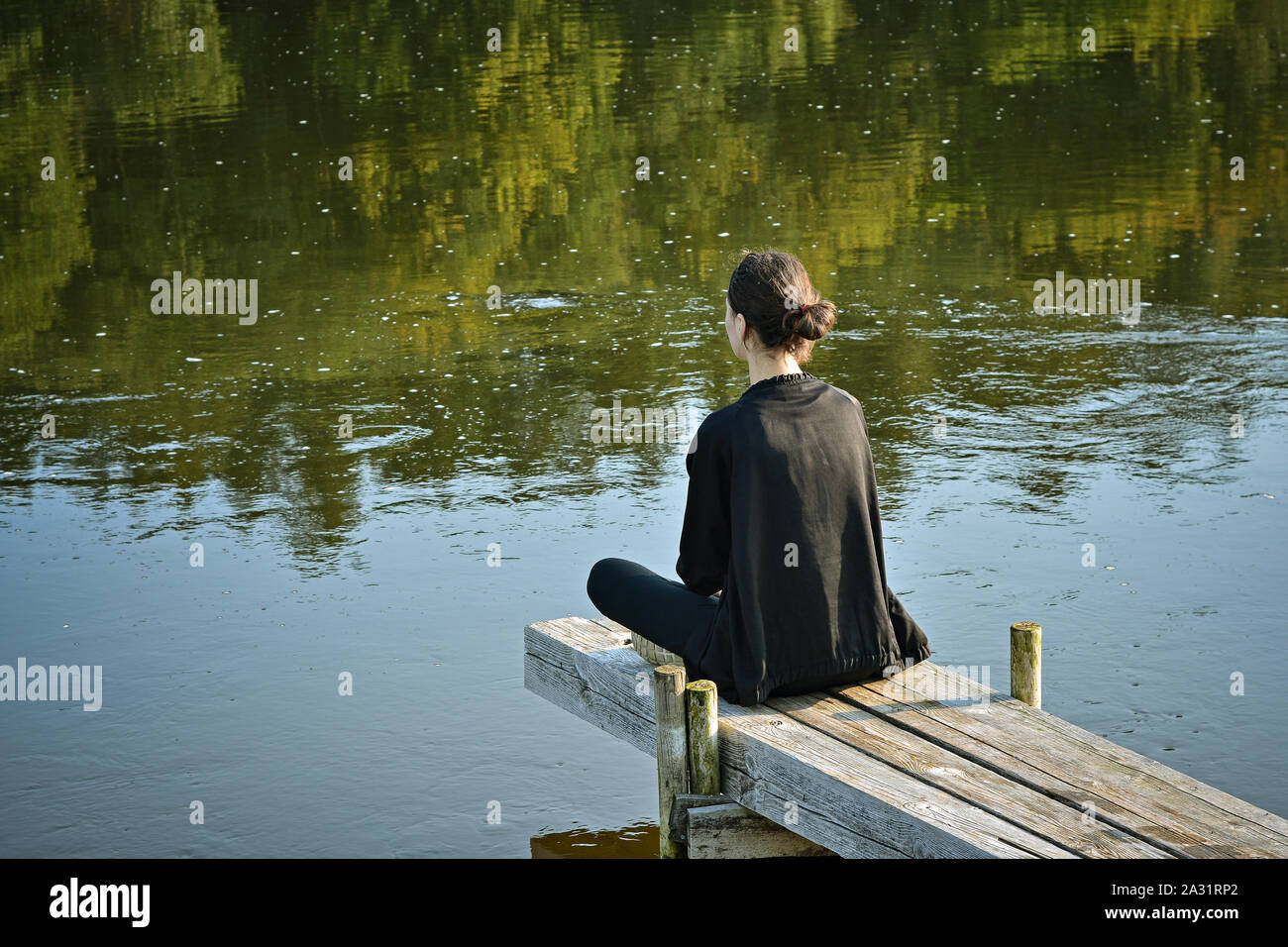 Junge Mädchen in einem Trainingsanzug und eine schwarze Jacke sitzt im Schneidersitz auf Gehwege und hält eine Meditation über die Wasseroberfläche des Flusses. Stockfoto