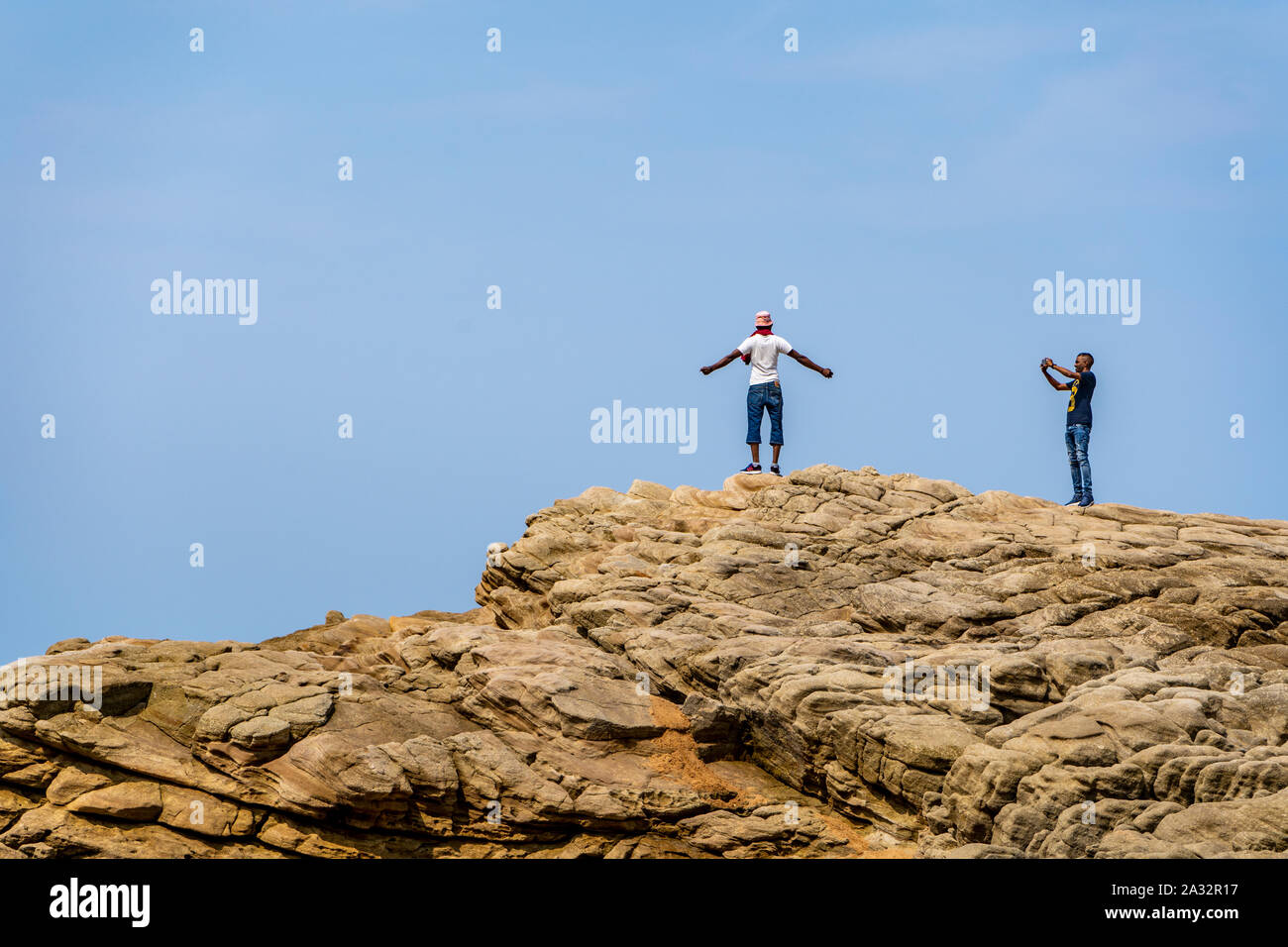 Durban, Südafrika - 21. September 2019: Der junge Mann stand an der Spitze der Rock mit ausgestreckten Armen auf den Strand als sein Freund nimmt sein Foto. Stockfoto