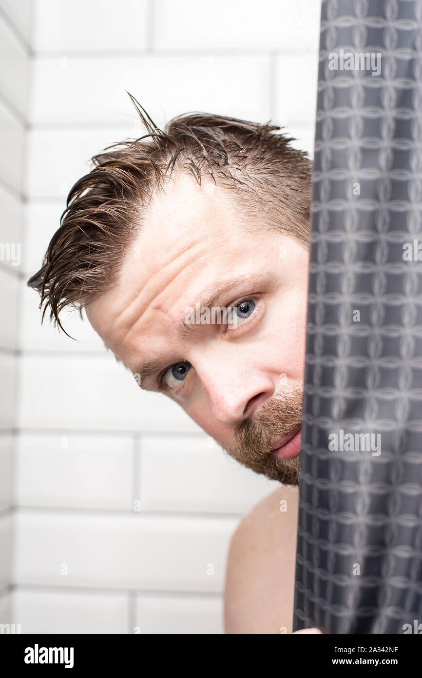 Gut aussehender, bärtiger Mann mit nassen Haar sieht aus hinter einem Vorhang in der Dusche. Close-up. Stockfoto