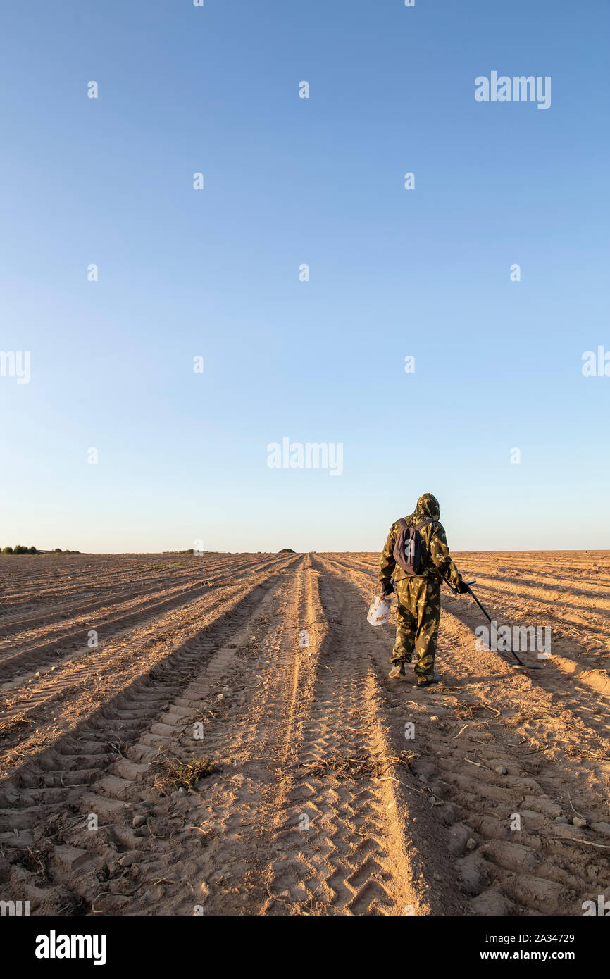 Ein Mann in der Tarnung ist auf der Suche nach Schatz mit einem Metalldetektor, im Feld, an einem Sommerabend bei Sonnenuntergang. Stockfoto