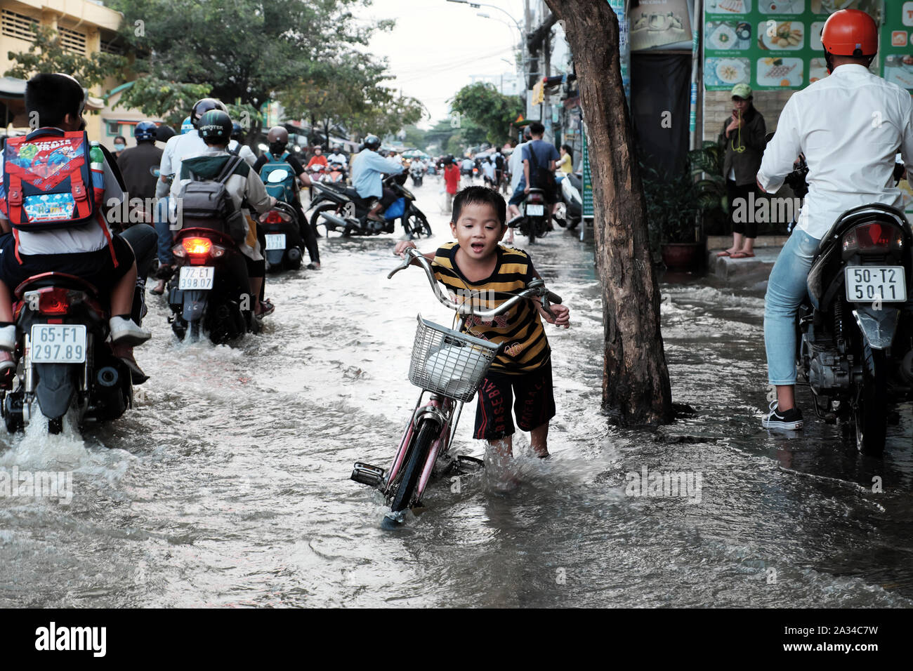 HO CHI MINH STADT, VIET NAM, kleiner Junge schieben Fahrrad überqueren überflutete Straße, schwer im Wasser durch Flut am Abend zu bewegen, Vietnam Stockfoto