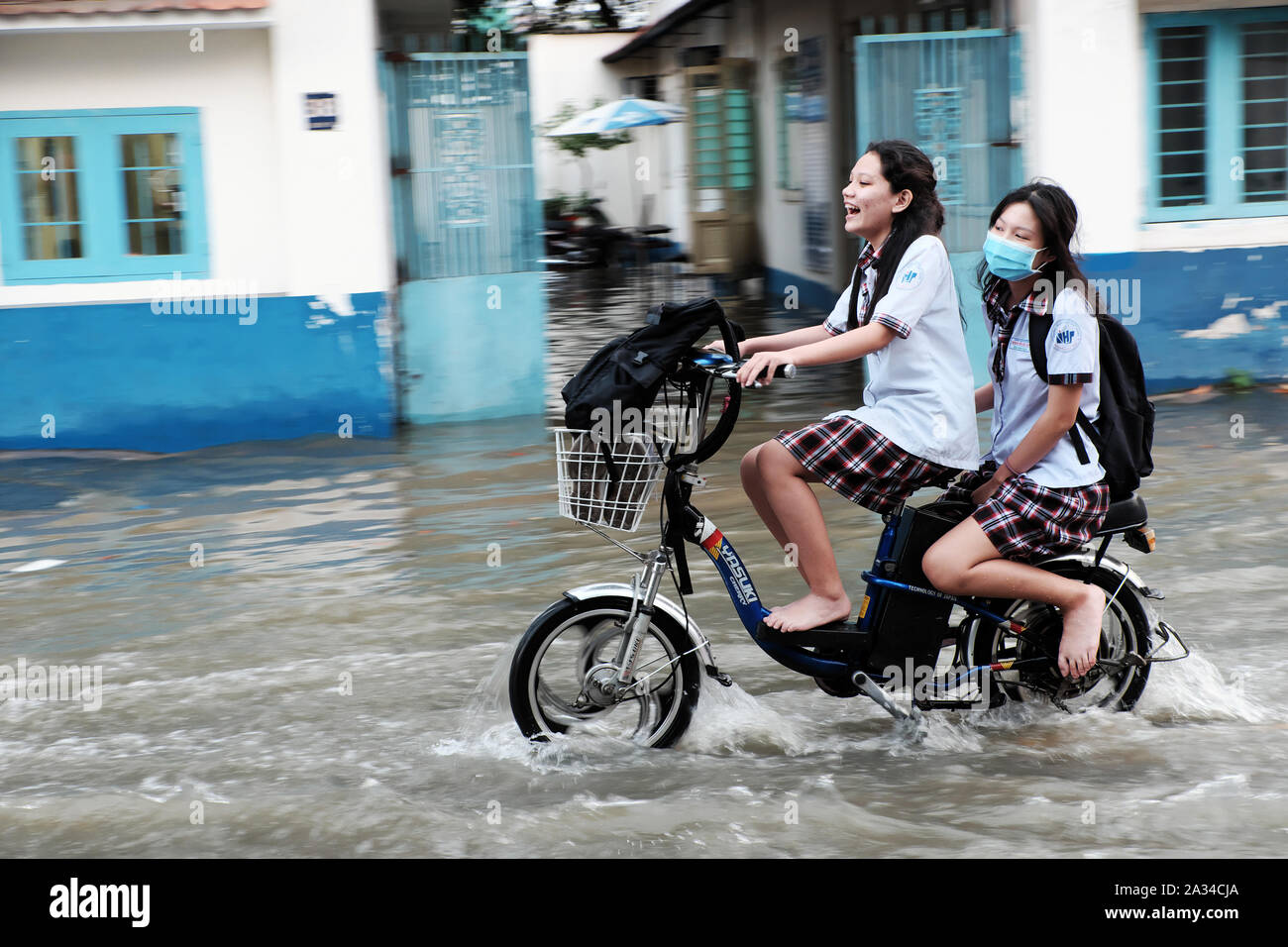 HO CHI MINH CITY, VIETNAM - Sept 30, 2019: 2 Schule Mädchen fahrt Elektrofahrrad Kreuzung überschwemmte Straße bei Flut am Abend, jugendlich Mädchen lachen, wenn Mo Stockfoto