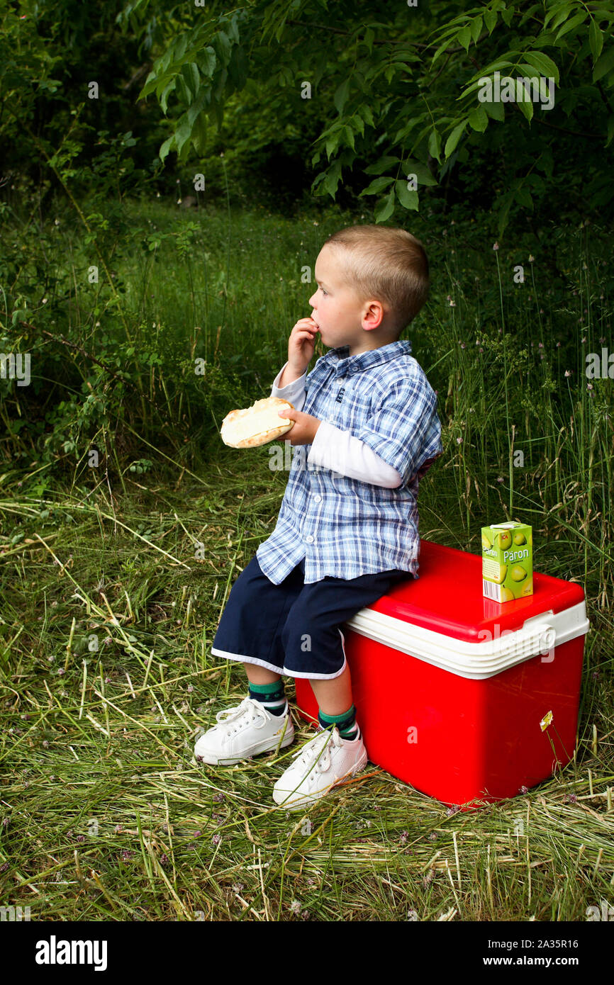 Junge sitzt auf Kühltasche und isst ein Sandwich Stockfoto