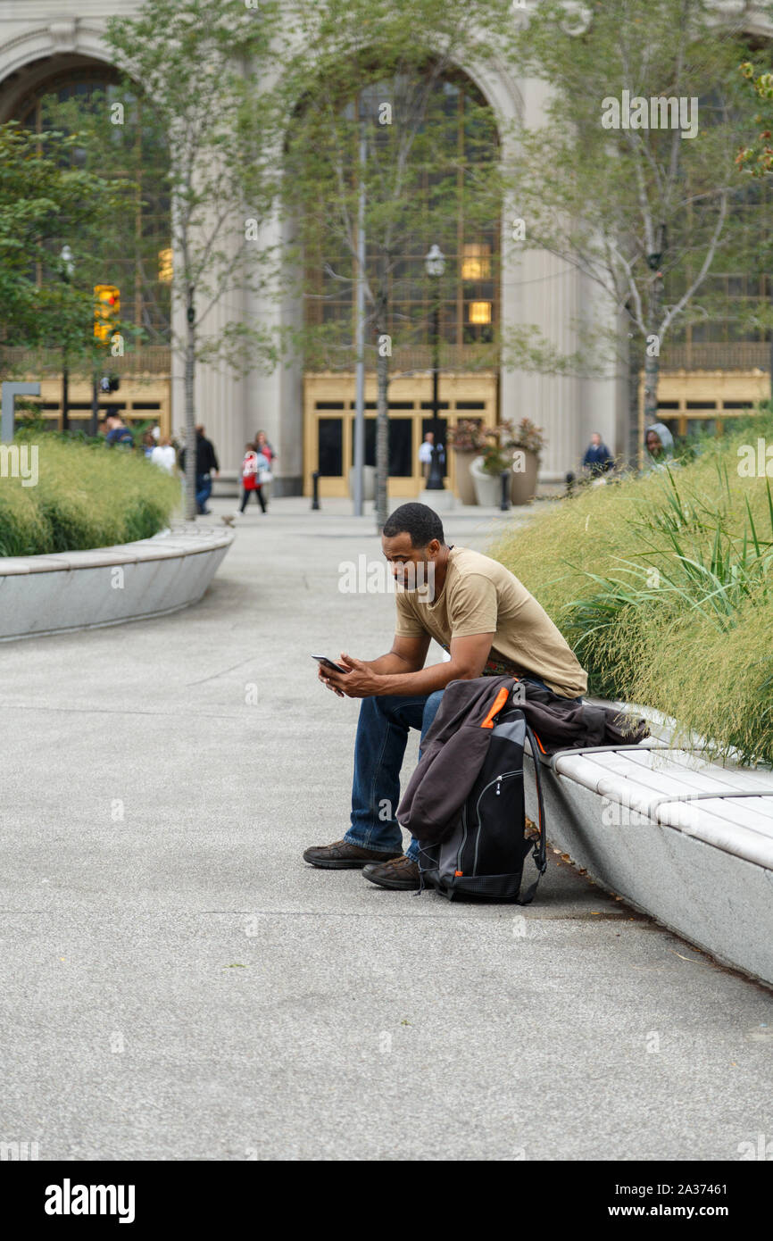 Eine sitzende Mann in casual Tuch schaut auf sein Handy an der Cleveland öffentlichen Platz mit Blick auf den Tower City Center. Cleveland, Ohio, USA. Stockfoto