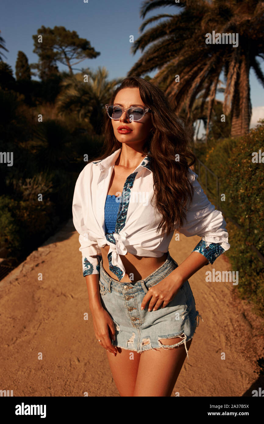 Schöne stilvolle Frau wandern in tropischen Park mit Palmen an einem sonnigen Abend. Braungebrannte Mädchen in Jeans Rock, Spitze blau, weiße Bluse und Sonnenbrille. Rel Stockfoto