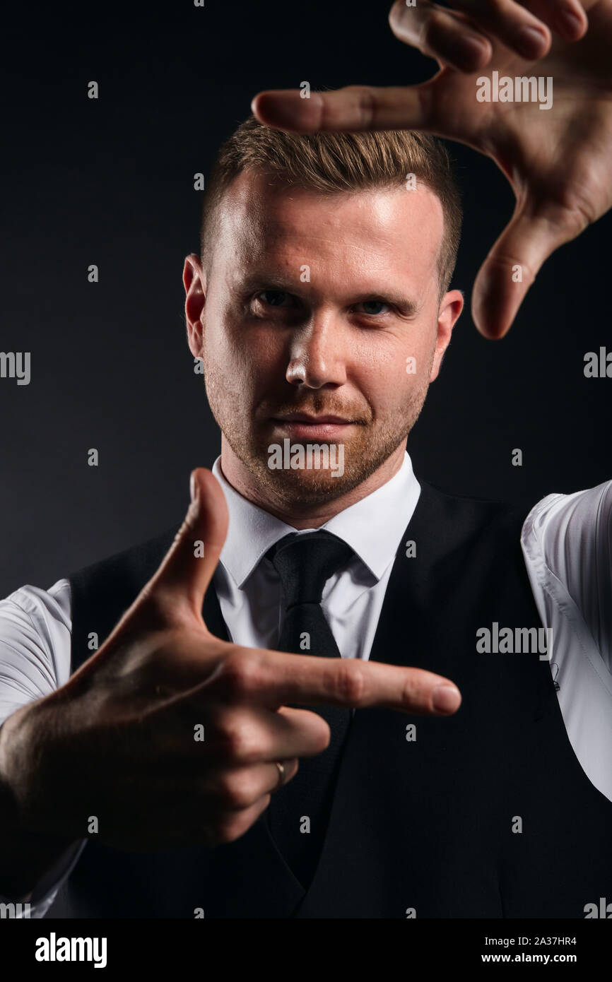 Jungen gutaussehenden Mann im weißen Hemd, schwarze Weste, Rahmen mit seinen Händen auf schwarzem Hintergrund, bis Foto schliessen. Isolierte schwarze Hintergrund Stockfoto