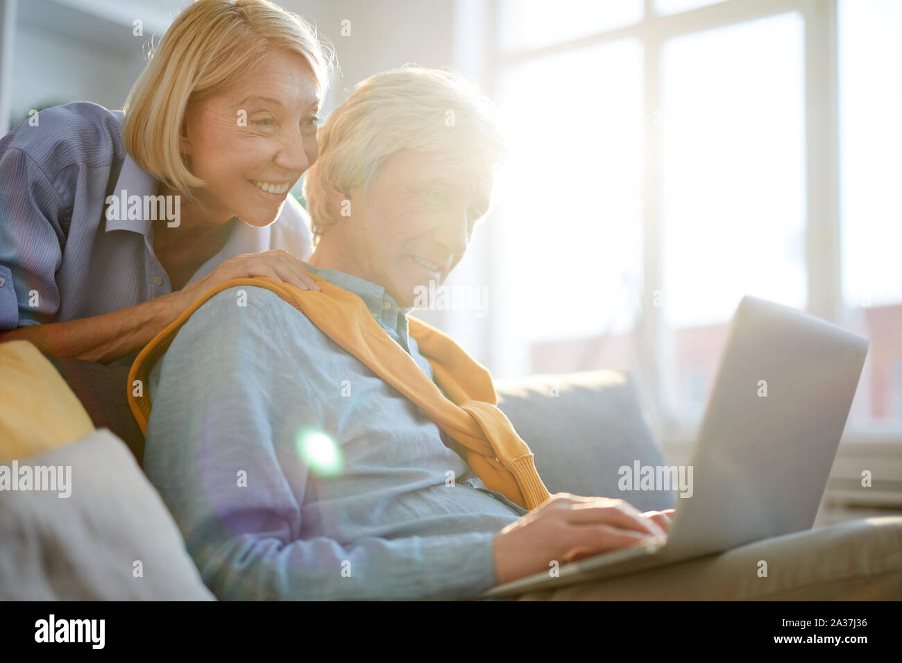 Portrait von älteren Mann mit Laptop, während die von zu Hause aus arbeiten mit fürsorglichen Frau ihn umarmen im Sonnenlicht, kopieren Raum Stockfoto