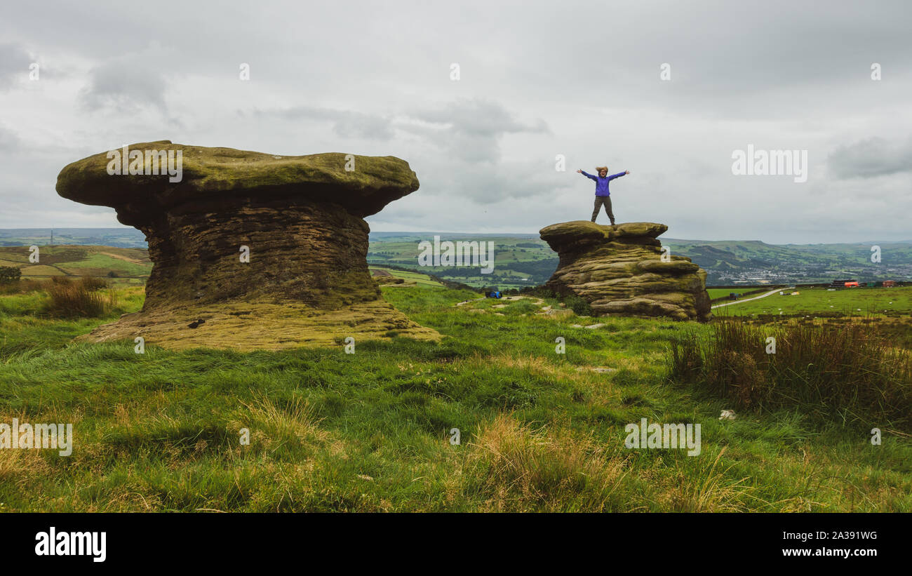 Person, die auf der Oberseite des Doppler Steine mit ausgestreckten Armen, Ilkley Moor, UK ihre ungewöhnliche Form, indem Sie die Oberseite der Festplatte grits verursacht wird Stockfoto