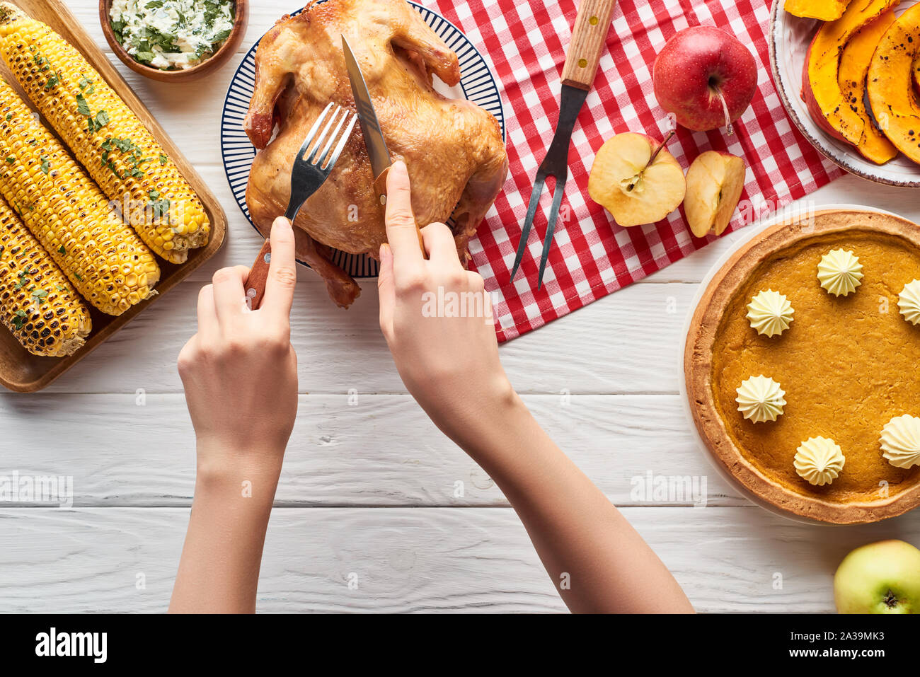 7/8-Ansicht von Frau schneiden gebratener Truthahn in der Nähe von Pumpkin Pie und gegrilltem Gemüse serviert auf weiße Holztisch mit Rot karierte Servietten Stockfoto