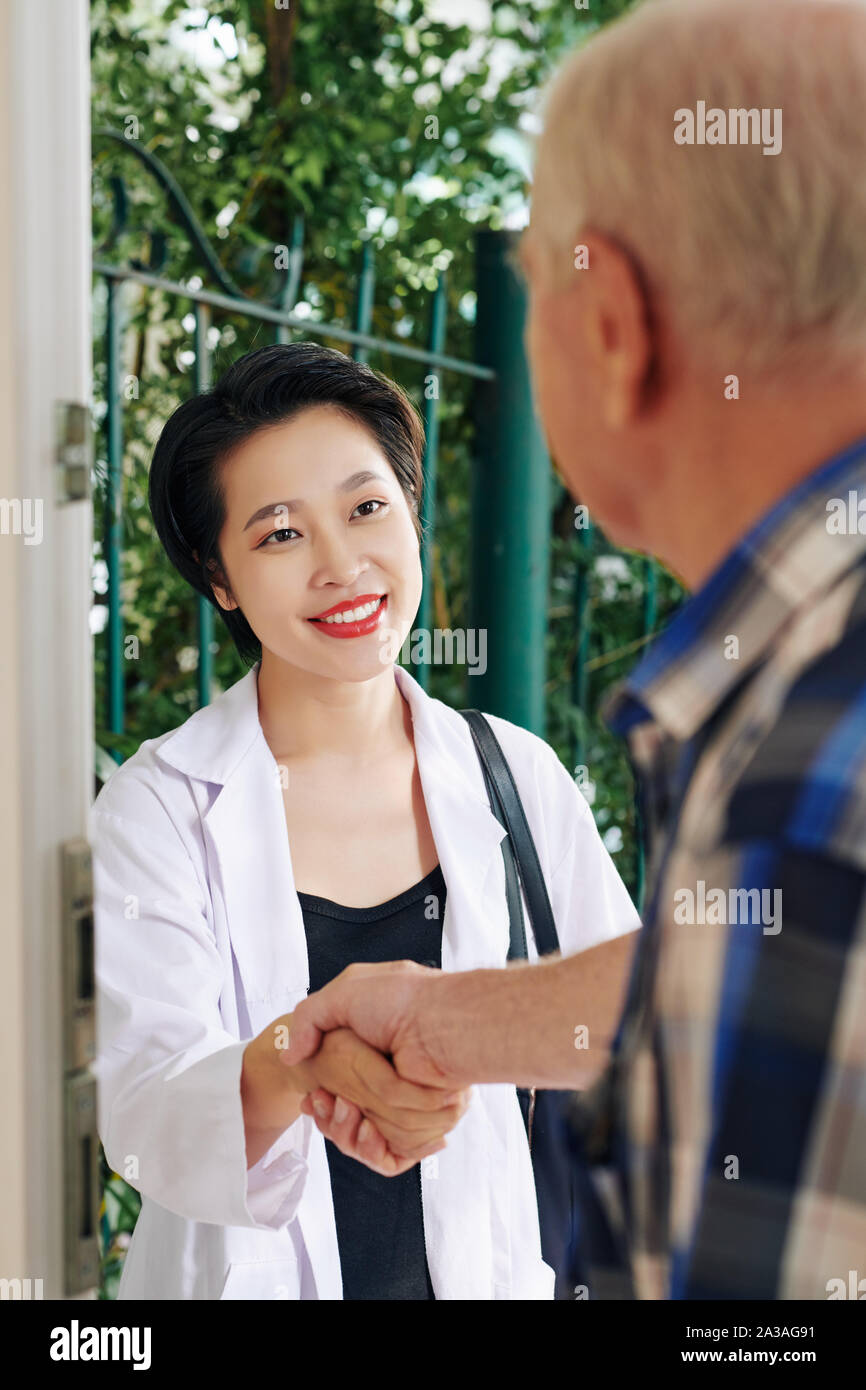 Ziemlich lächelnden jungen vietnamesischen Arzt zitternden Hand des älteren Menschen nach ihm zu Hause besuchen und diskutieren Therapie Prozess Stockfoto
