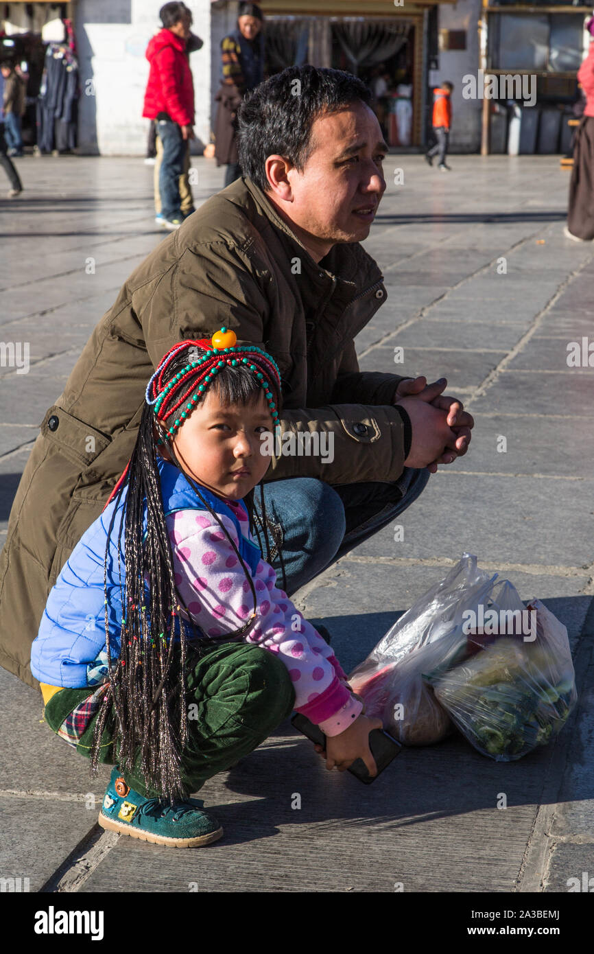 Eine junge tibetische Mädchen, Sitzen, die von ihrem Vater, trägt Eine Perücke der Khamba Stil Zöpfe und eine rote Koralle Kopfschmuck in Barkhor in Lhasa, Tibet. Stockfoto