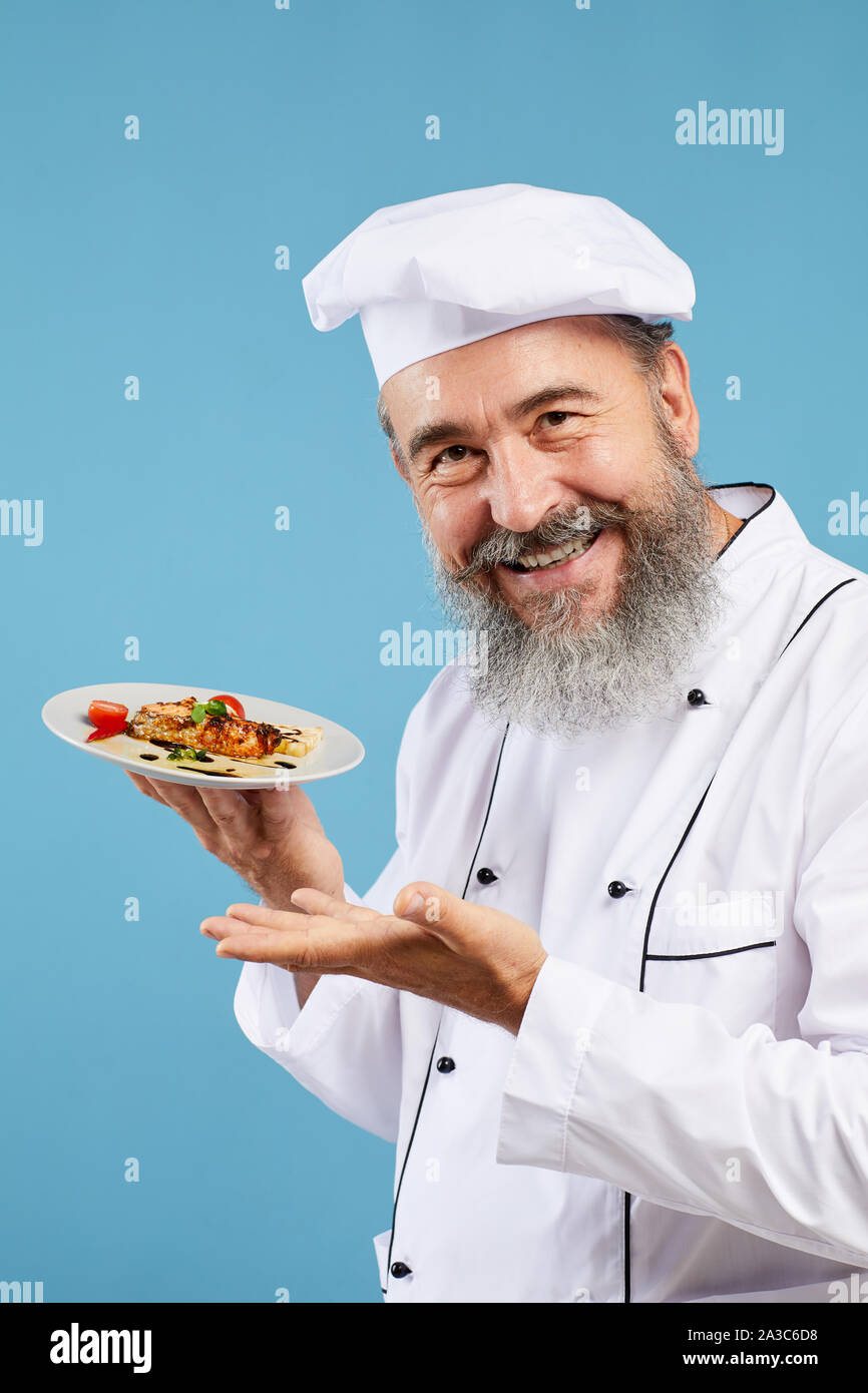 Portrait von fröhlichen Senior Chef präsentiert italienische Gericht beim Stehen vor blauem Hintergrund und lächelnd an Kamera Stockfoto