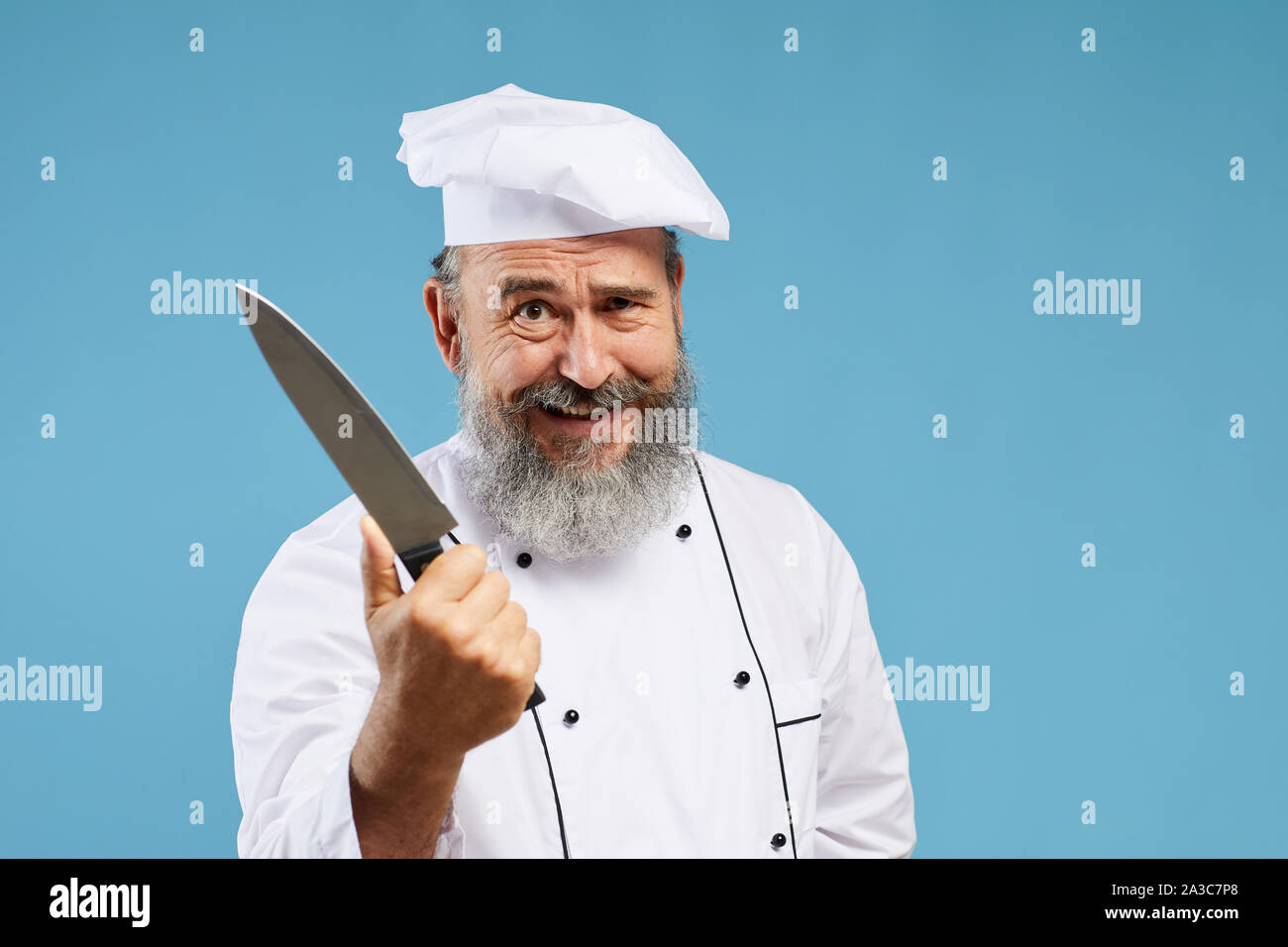 Portrait von verrückt aussehenden bärtigen Koch Holding scharfen Messer und lächelnd an der Kamera beim Stehen vor blauem Hintergrund, kopieren Raum Stockfoto