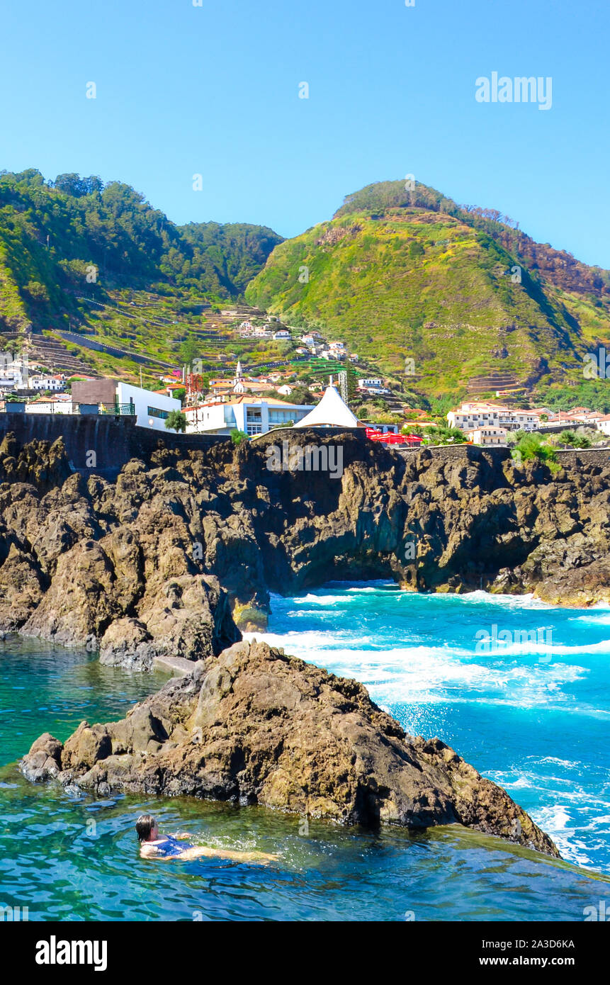 Lady Schwimmen in natürlichen Pools im Atlantischen Ozean, Insel Madeira, Portugal. Aus vulkanischem Gestein, in das Meer natürlich fließt. Stadt Porto Moniz auf dem Berg im Hintergrund. Stockfoto