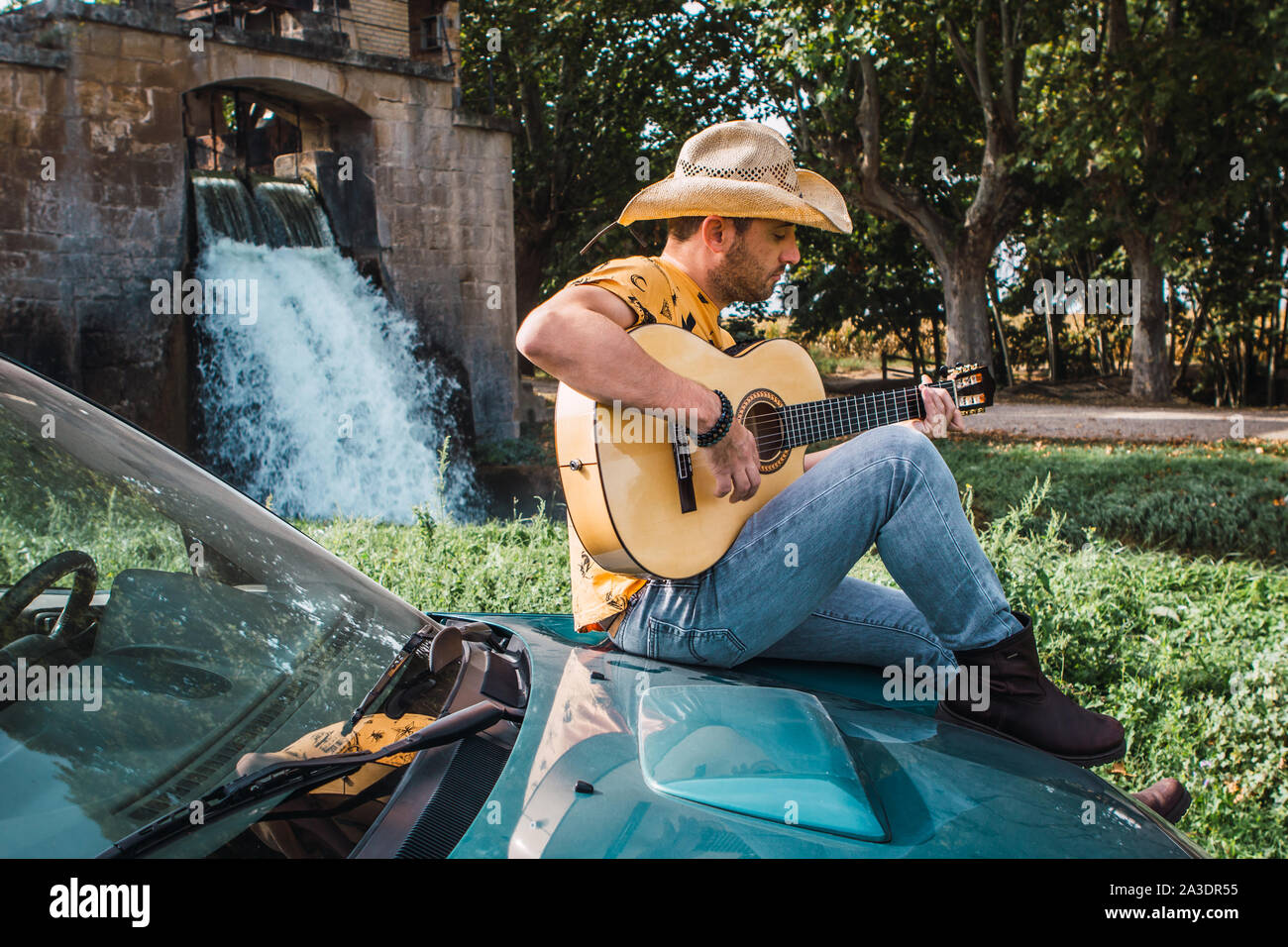 Mittleres alter Mann mit einem Cowboyhut auf ein geländegängiges Fahrzeug während des Spielens der Gitarre im Freien Stockfoto