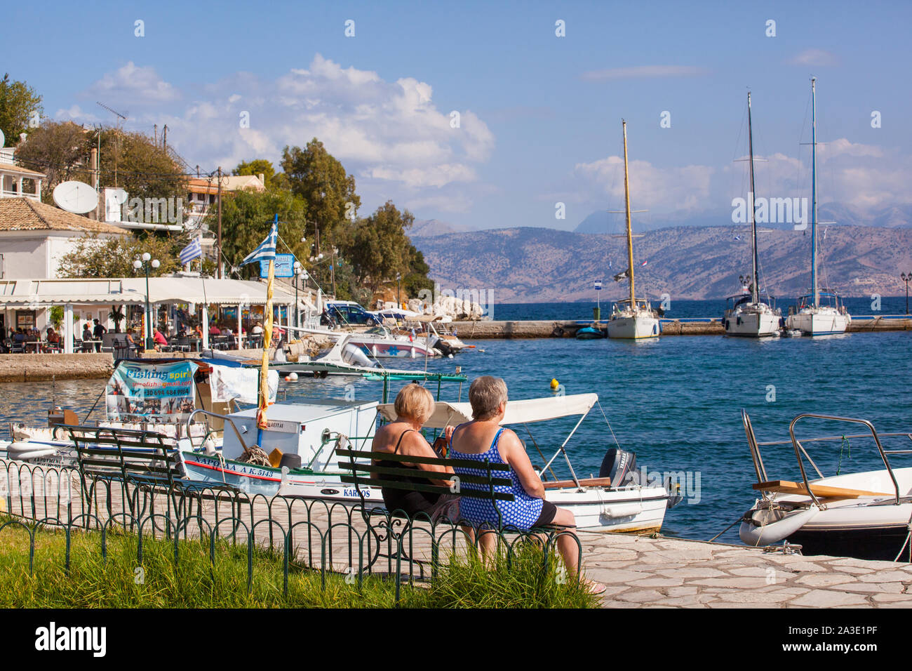 Zwei Frauen Urlauber durch den Hafen in der Griechischen Ferienort Kassiopi Korfu Griechenland sitzen mit Booten in See und blauer Himmel Himmel Stockfoto