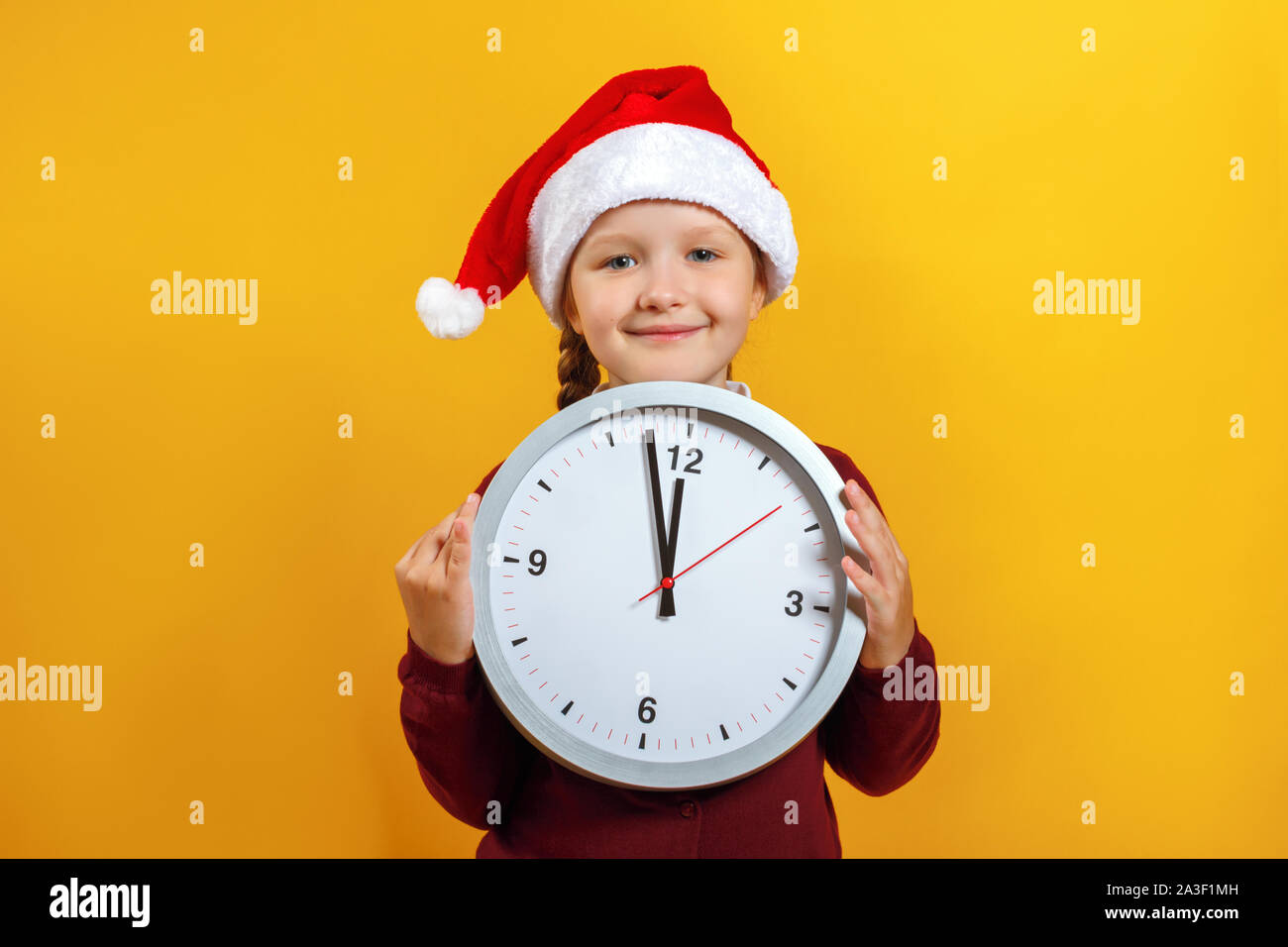Heiligabend. Portrait Of Happy charmante kleine süße Mädchen in Santa's Hut. Das Kind hält eine Uhr auf einem gelben Hintergrund. Stockfoto