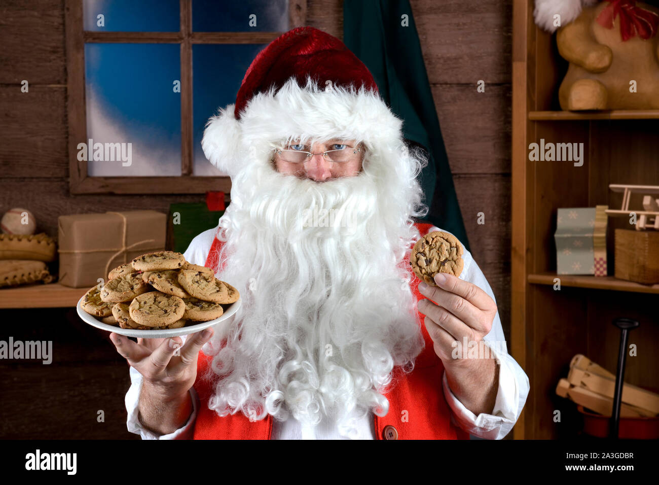 Santa Claus in seiner Werkstatt mit einem Teller voll von frisch gebackenen Chocolate Chip Cookies in der einen Hand und einem Keks in der anderen. Stockfoto