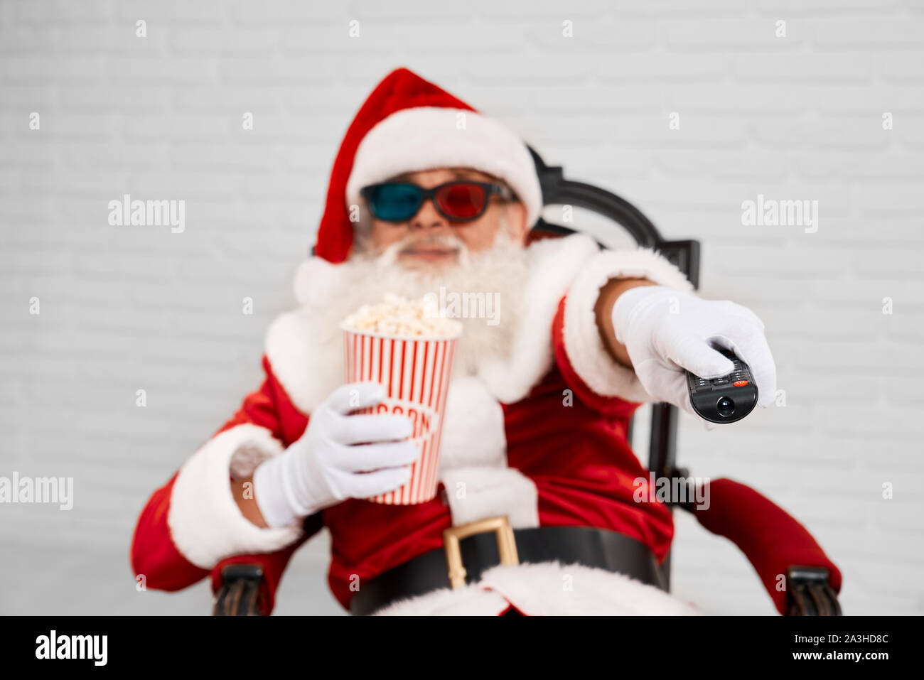 Portrait von Sankt Nikolaus in Rot und Weiß Kostüm im Sessel sitzen,  fernsehen in 3D-Brille und mit Zapper tv zu wechseln. Ältere Mann mit  grauem Bart essen Popcorn und genießen Film Stockfotografie -
