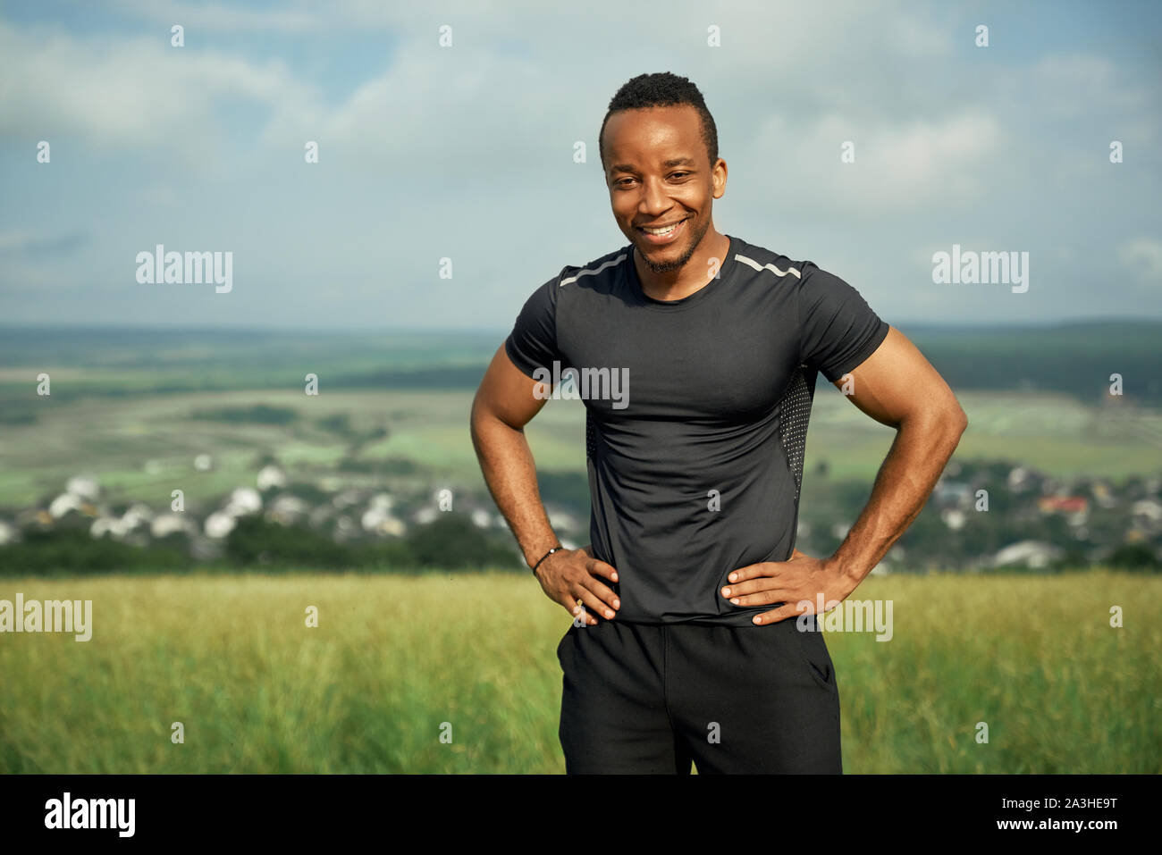 Portrait von lächelnden, stattlich Sportler im Freien auf den Hüften. Muskulöse Sportler tragen in schwarzen T-Shirt und Shorts. Positive Athlet auf dem Hintergrund der Natur und blauer Himmel posieren. Stockfoto